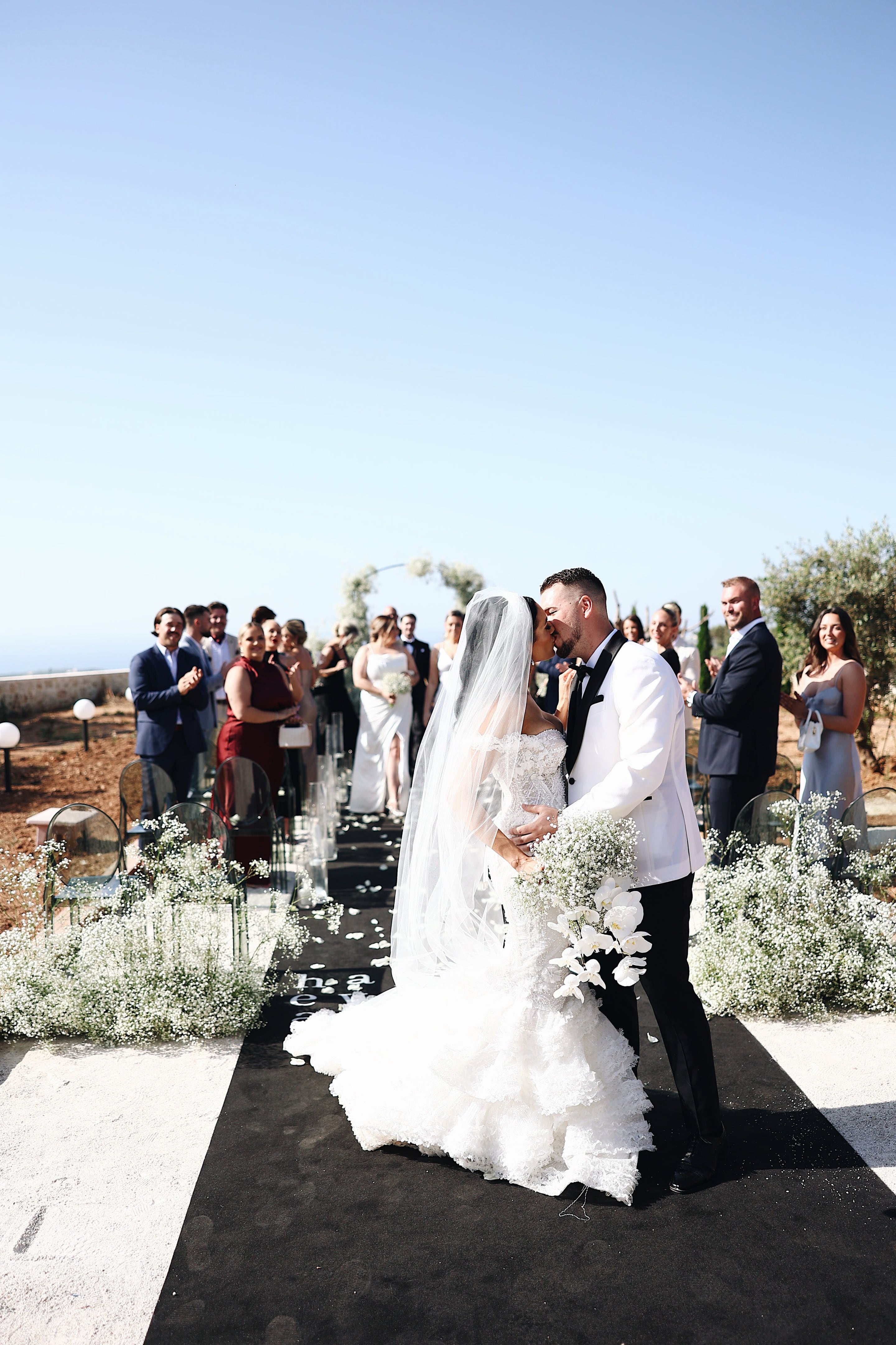 A bride in a tiered, lace-textured gown and long veil kisses her groom in a white dinner jacket and black tie on a black aisle runner strewn with petals, as guests applaud beneath an arch of white blooms. Frothy baby's breath spills along both sides of the aisle, and the groom holds a bouquet of white orchids, at Villa Amnalys in Greece, a wedding ceremony in bright, open sunshine.