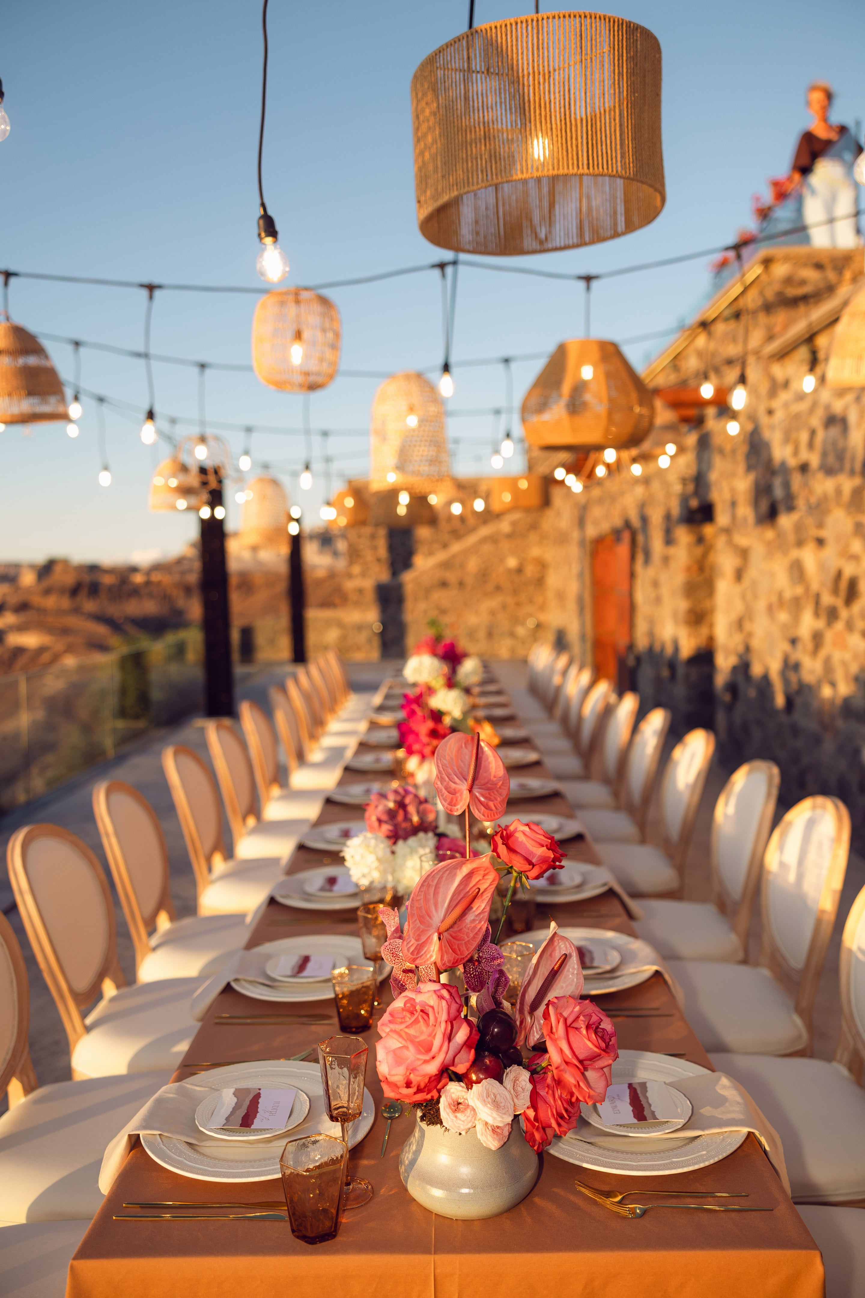 A long wedding dining table on a stone terrace at Cavo Ventus in Greece, lined with cream chairs and set with white plates and amber glassware. Down the centre sit low arrangements of coral anthurium, deep pink roses, white hydrangea and pale garden roses, with trailing orchids and dark berries. Warm evening light falls across the stone walls, and string lights and woven lanterns hang overhead above the view.