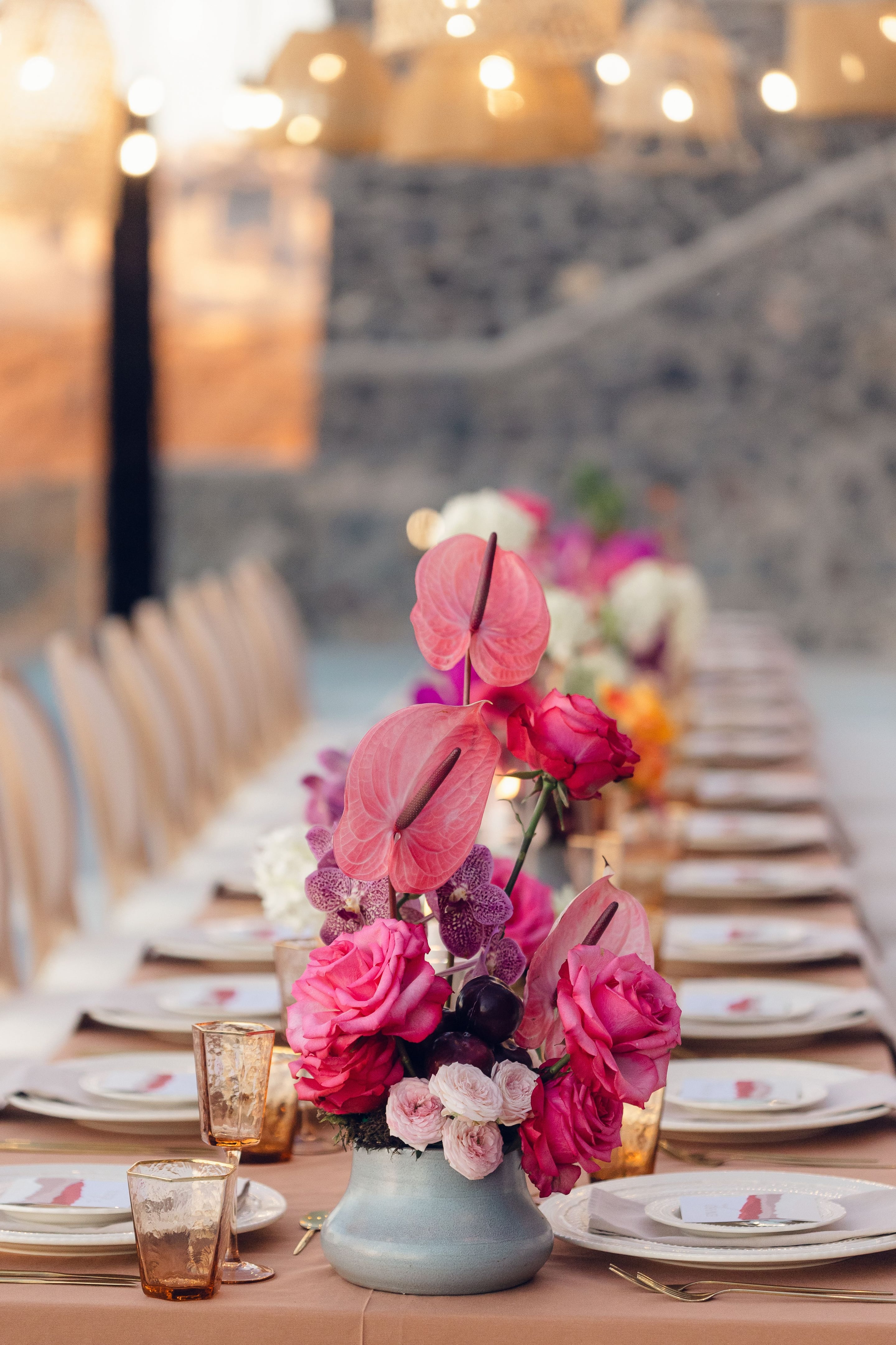 A low centrepiece in a pale blue-grey ceramic vase on a long table, with hot pink garden roses, pink anthurium blooms on tall stems, speckled purple orchids, dark plums and small blush roses. It is one of many similar arrangements down a wedding table at Cavo Ventus in Greece, with amber glasses and white place settings in the soft evening light.