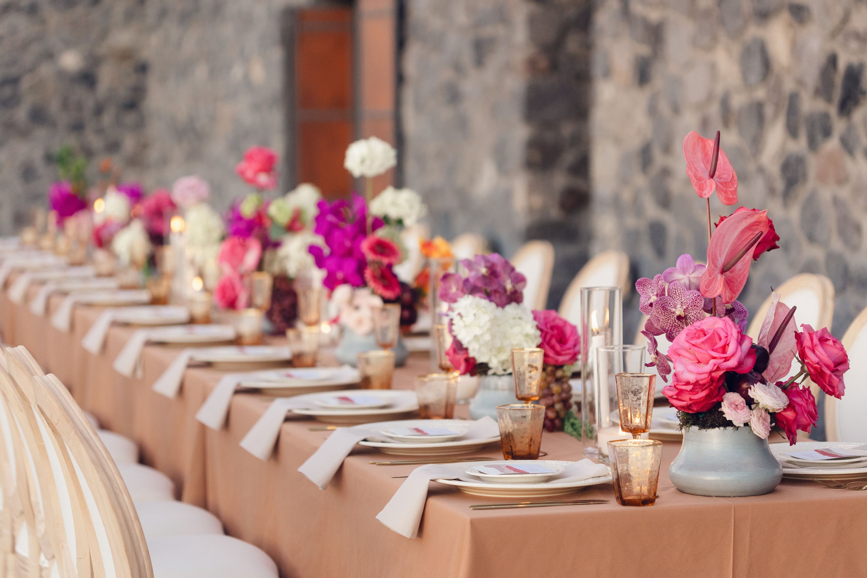 A long wedding banquet table dressed in a warm peach cloth at Cavo Ventus in Greece, lined with low centrepieces of hot pink garden roses, pink anthurium, magenta and lilac vanda orchids, and white hydrangeas in pale blue-grey vessels. Amber mercury glass goblets, white napkins and slim candle hurricanes sit beside each place setting, while the rough grey stone wall behind softens the vivid colours into an intimate evening mood.