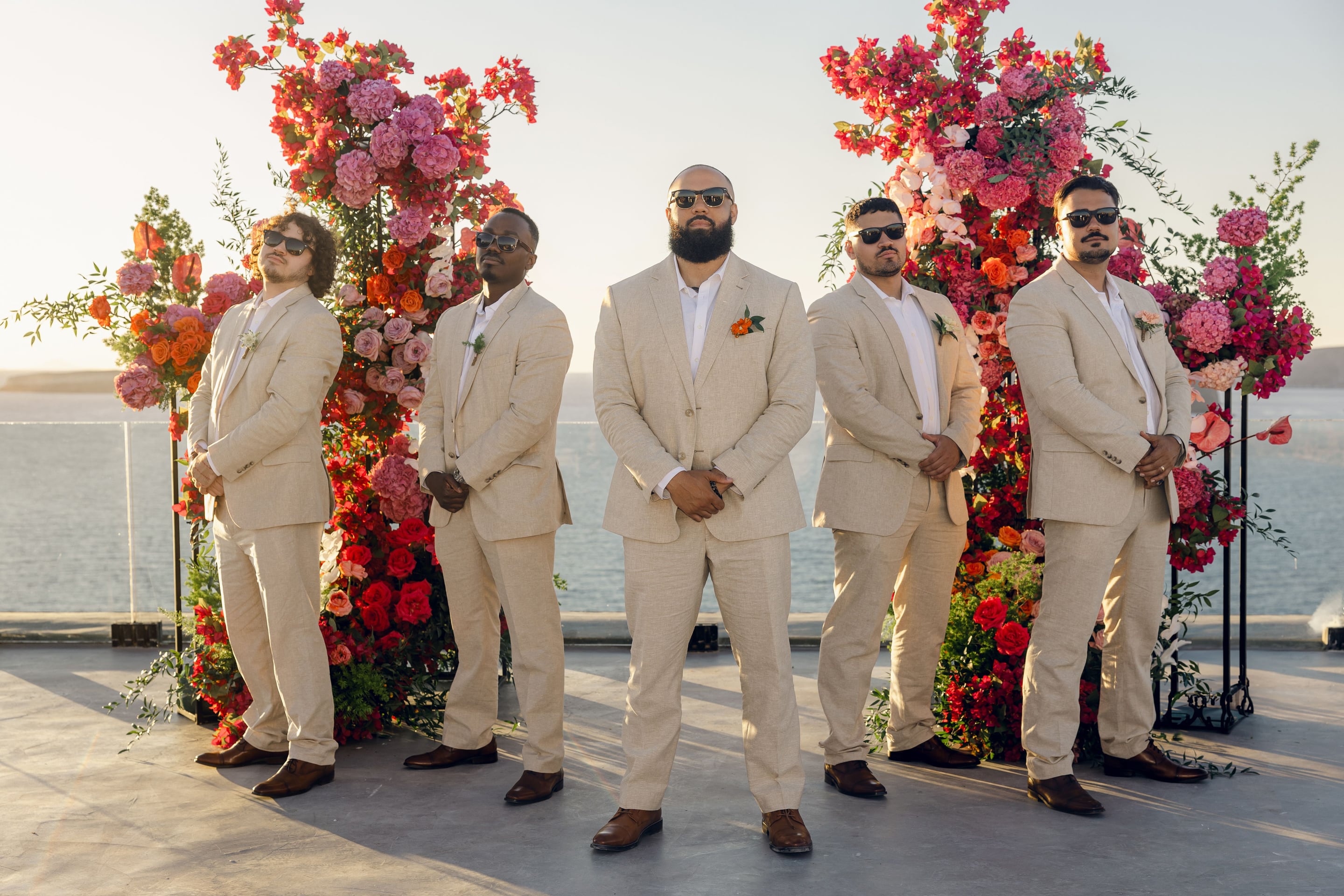 Five men in matching cream linen suits and white shirts stand in a row on a stone terrace, most wearing dark sunglasses, in front of a tall wedding arch heaped with bougainvillea, pink hydrangeas, coral roses and orange blooms. The sea glows pale blue behind them under a soft evening sky, part of the wedding flowers at Cavo Ventus in Greece. The central groom has a small orange flower in his lapel.