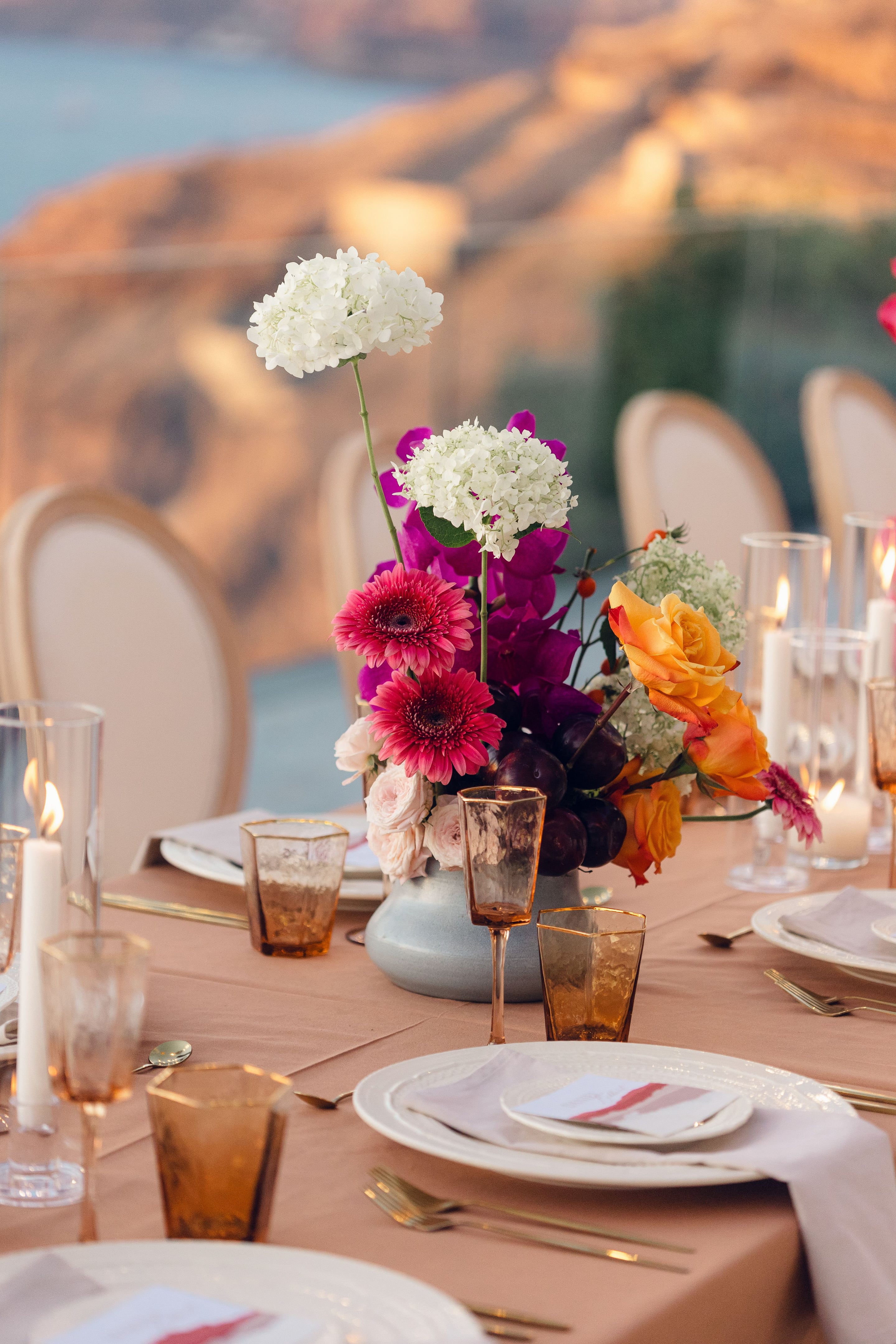 A wedding table centrepiece in a squat grey-blue ceramic vase, crowded with hot pink gerbera daisies, magenta orchids, orange roses, white hydrangea and blush garden roses, with dark plums tucked among the stems. It sits on a dusty rose tablecloth at Cavo Ventus in Greece, flanked by amber glasses, gold cutlery, white plates and tall pillar candles in glass hurricanes.