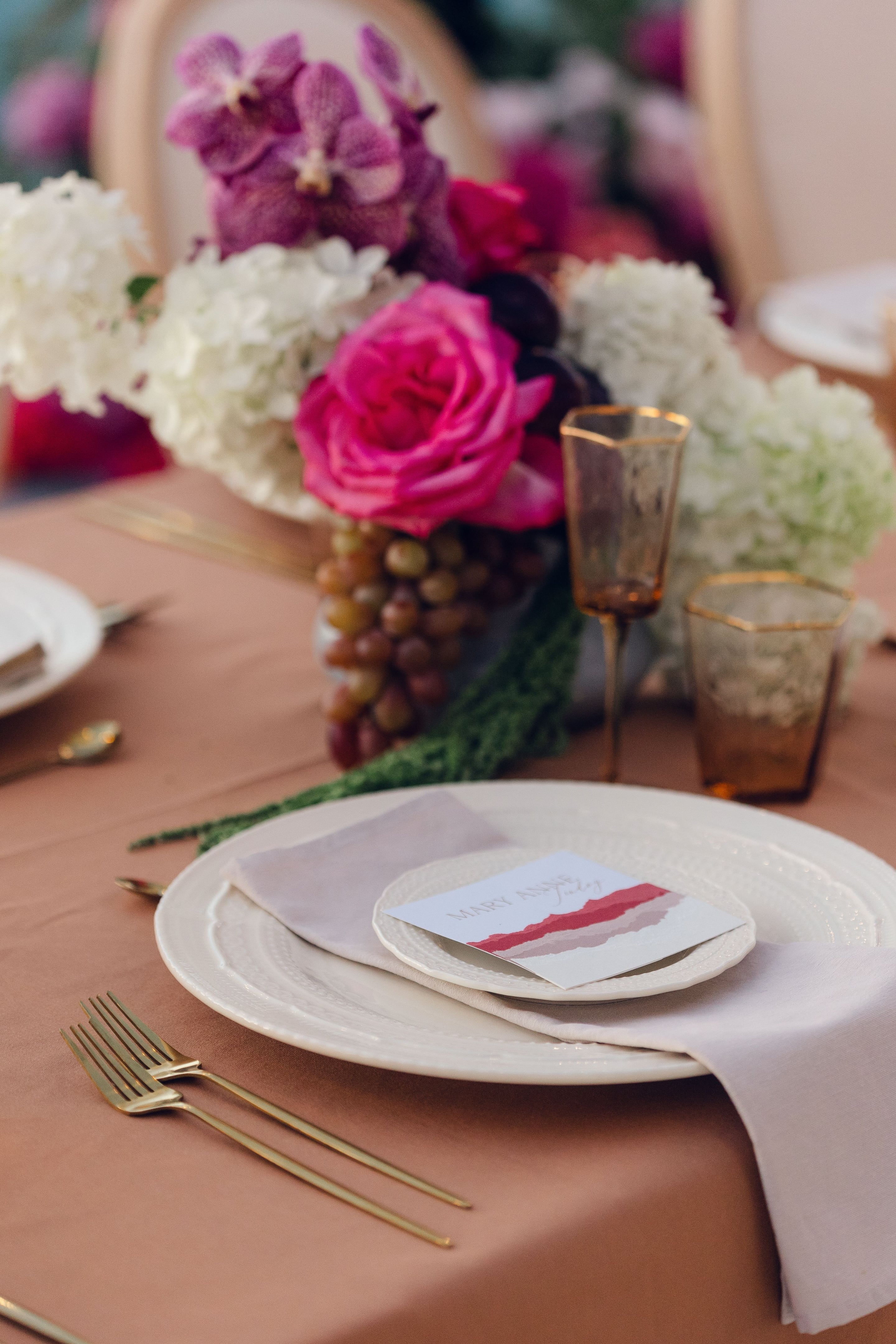 A wedding table setting at Cavo Ventus in Greece, where a centrepiece of hot pink garden roses and white hydrangeas rises above clusters of green and purplish grapes. Trailing green foliage runs across a dusty pink tablecloth, beside amber-tinted glasses with gold rims and a white plate holding a card with a painted pink and mauve stripe. The soft, warm light gives the table a romantic, elegant mood.