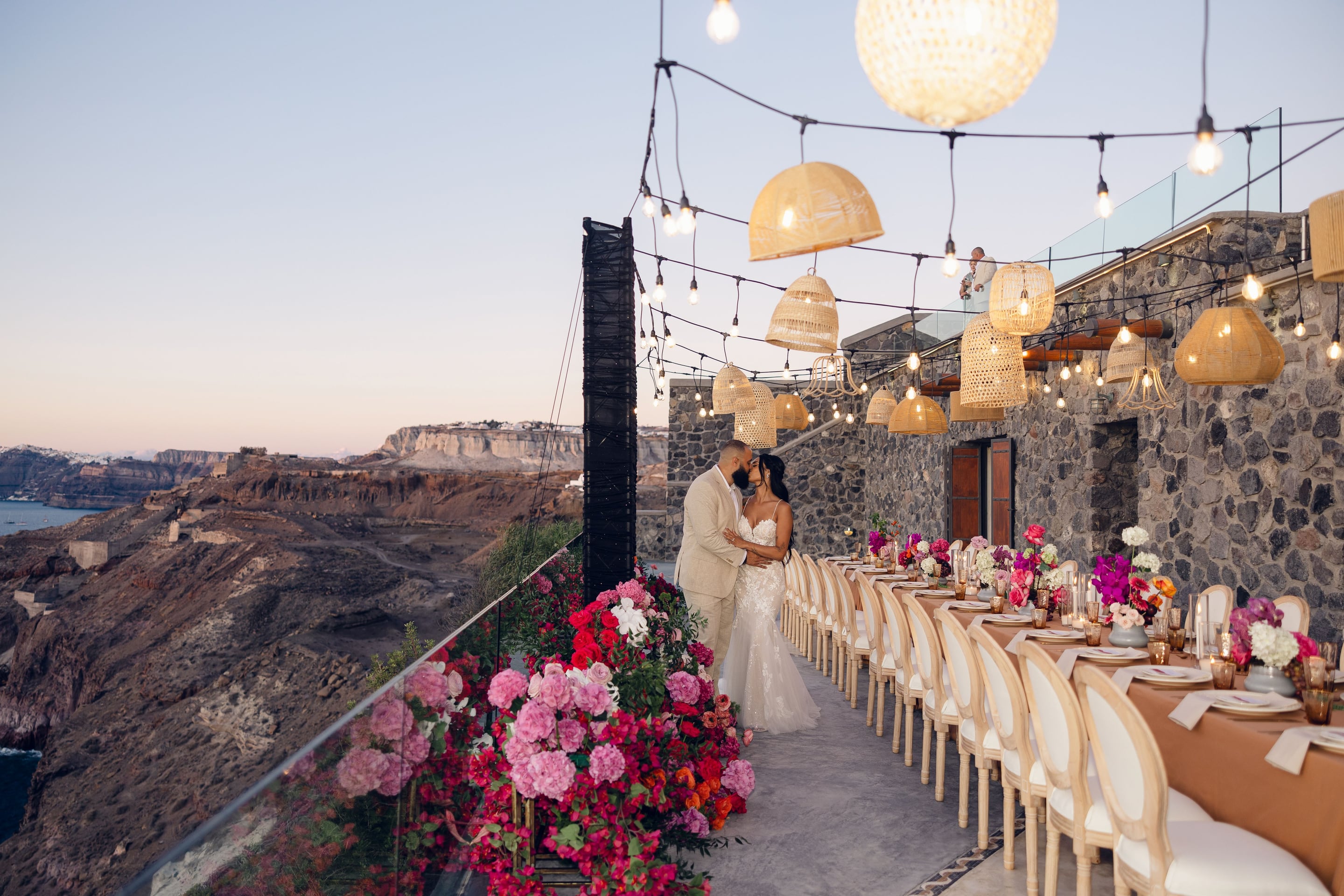 A bride in a fitted lace gown and a groom in a pale linen suit share a kiss at dusk on a stone terrace at Cavo Ventus in Santorini, Greece, beside a long wedding dining table. Pink hydrangeas, fuchsia bougainvillea and red roses spill along the glass railing in a lush border, while the table is dressed in terracotta linen with cream chairs and low, mixed flower centrepieces. Rattan pendant lamps and strings of bulbs glow overhead, and the caldera cliffs fade into a soft lavender sky behind them.