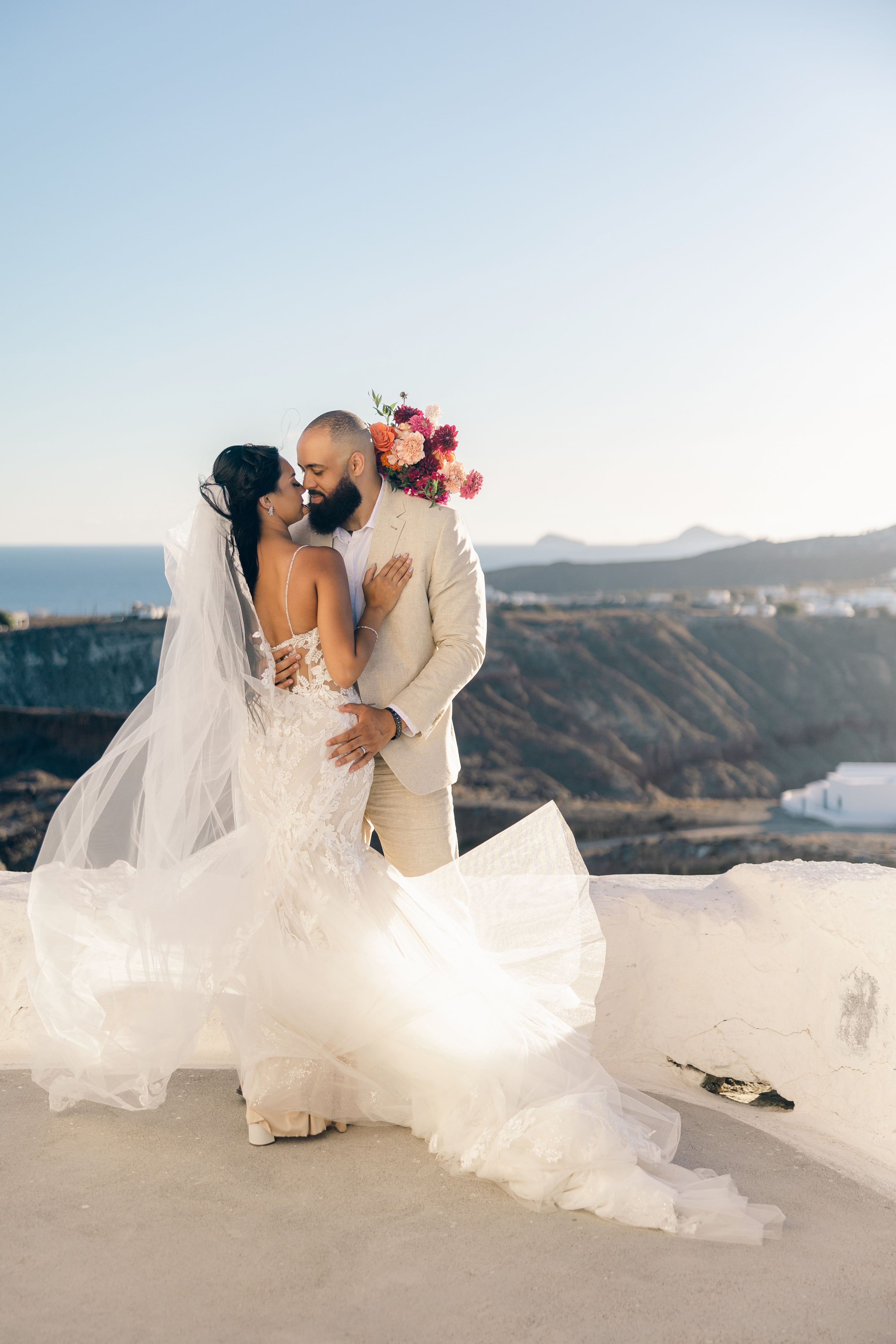 A bride in a fitted lace gown with a sheer open back and a long veil lifting in the breeze leans in to kiss a bearded groom in a cream linen suit, while he holds her waist. He carries a bridal bouquet of coral roses, pink ranunculus and deep magenta dahlias. Behind them, a cliffside terrace at Cavo Ventus in Santorini, Greece, opens onto a hazy blue sea and golden evening light, with a white-washed chapel nearby.