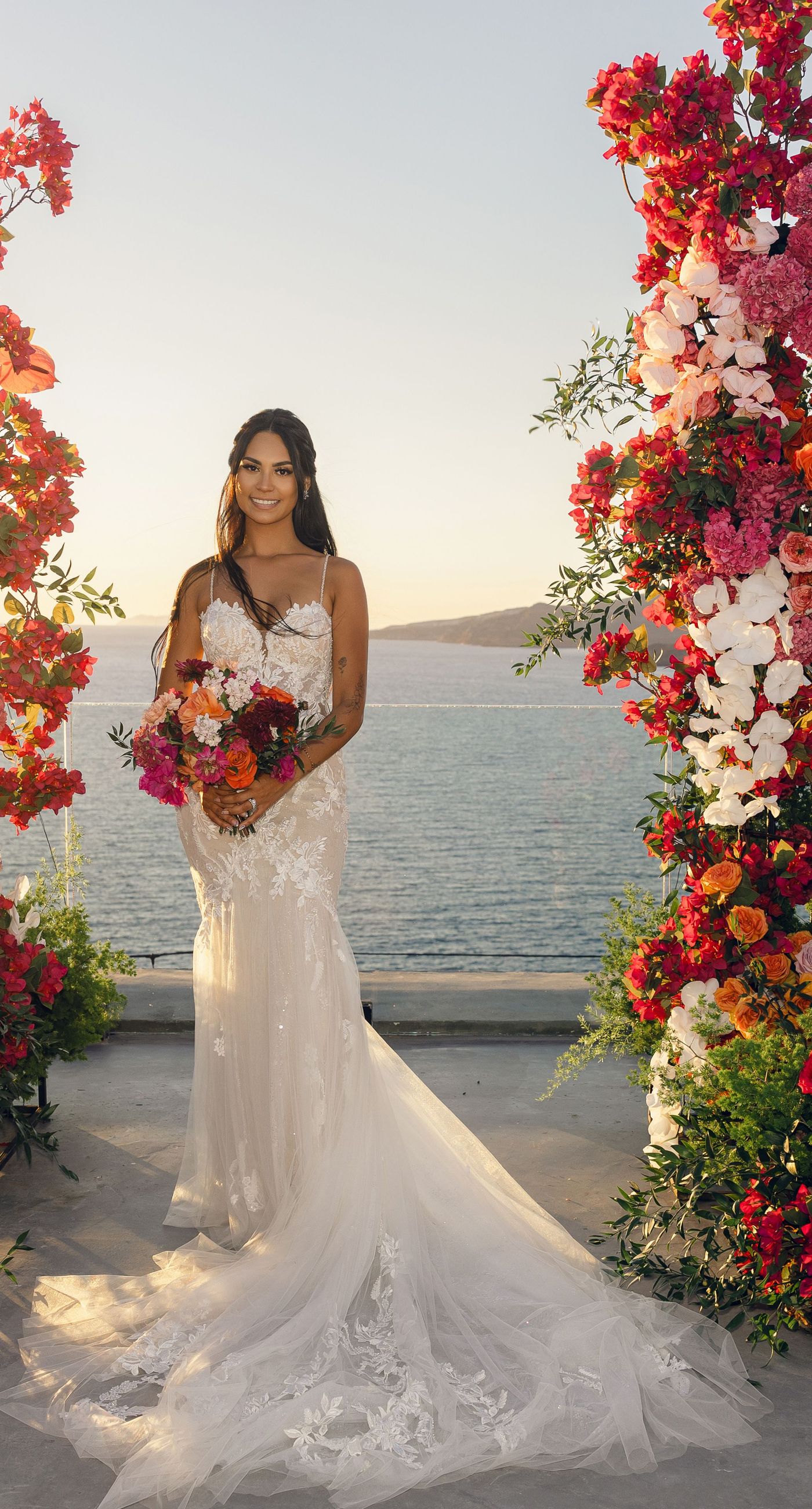 Chris & Jazmin - In a dreamlike setting beneath the soft embrace of the sun, this captivating work embodies a perfect union of elegance and romance. The delicate lace of her gown interlaces with the tender embrace of her partner, creating an ethereal atmosphere amidst the tranquil architecture. A vibrant bouquet bursts with life, featuring an exquisite medley of fuchsia, orange, and deep crimson blooms, each petal whispering tales of timeless love. The muted tones of the setting contrast beautifully with the vivid flowers, offering a glimpse into a serene world where artistry and nature harmonize flawlessly. This presentation speaks to the profound beauty that can blossom in moments of connection. - Photo 3