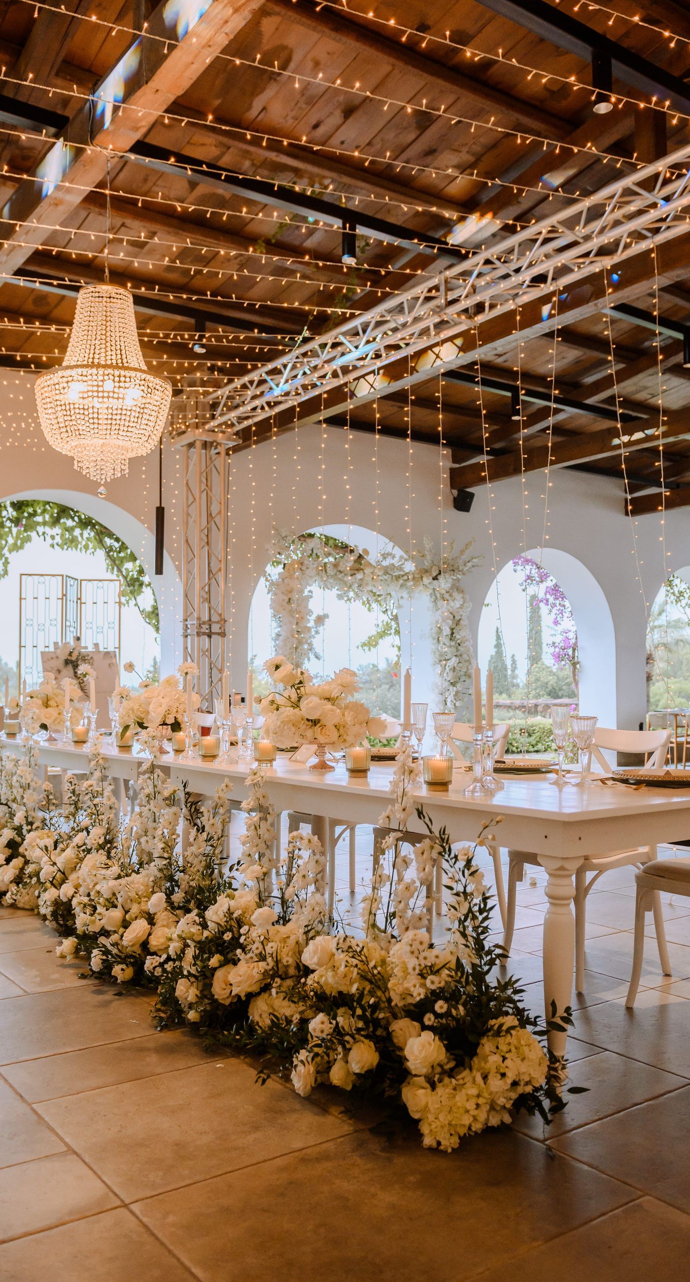 Christine & Eduart - Under a delicate floral arch, adorned with lush white blossoms and greenery, stands a vision of elegance and romance. The gentle pastel hues of the surrounding florals beautifully complement the soft, ethereal ambiance of this enchanting scene. It feels as though nature itself has been curated to frame a moment of serene connection, blending seamlessly into a verdant backdrop. The contrast of the bride's exquisite, flowing gown with the groom's tailored, modern attire in shades of emerald and white speaks volumes of both tradition and contemporary flair. This work captures an essence of timeless beauty, harmoniously blending natural elements with sophisticated design. - Photo 6