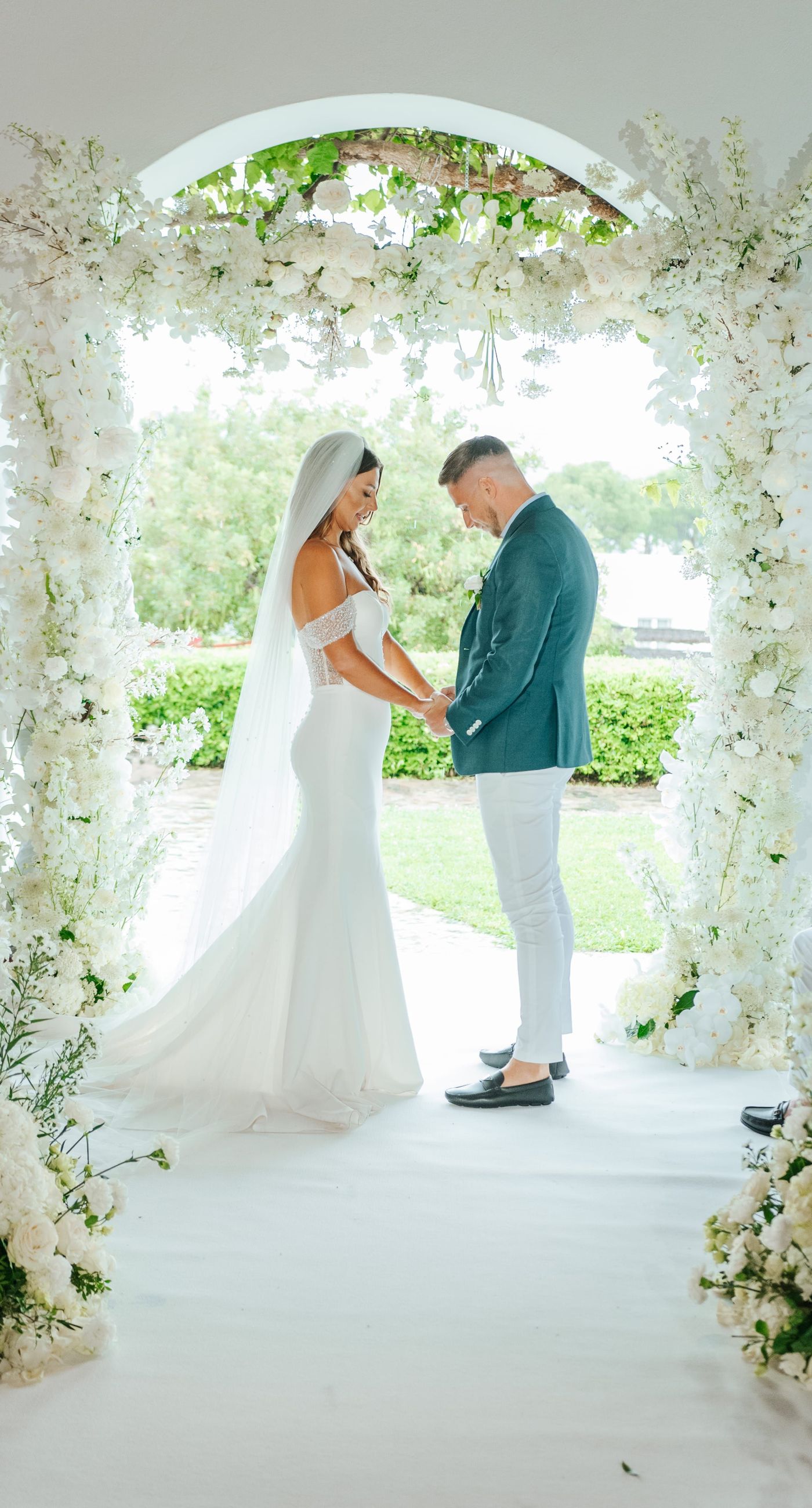 Christine & Eduart - Under a delicate floral arch, adorned with lush white blossoms and greenery, stands a vision of elegance and romance. The gentle pastel hues of the surrounding florals beautifully complement the soft, ethereal ambiance of this enchanting scene. It feels as though nature itself has been curated to frame a moment of serene connection, blending seamlessly into a verdant backdrop. The contrast of the bride's exquisite, flowing gown with the groom's tailored, modern attire in shades of emerald and white speaks volumes of both tradition and contemporary flair. This work captures an essence of timeless beauty, harmoniously blending natural elements with sophisticated design. - Photo 1