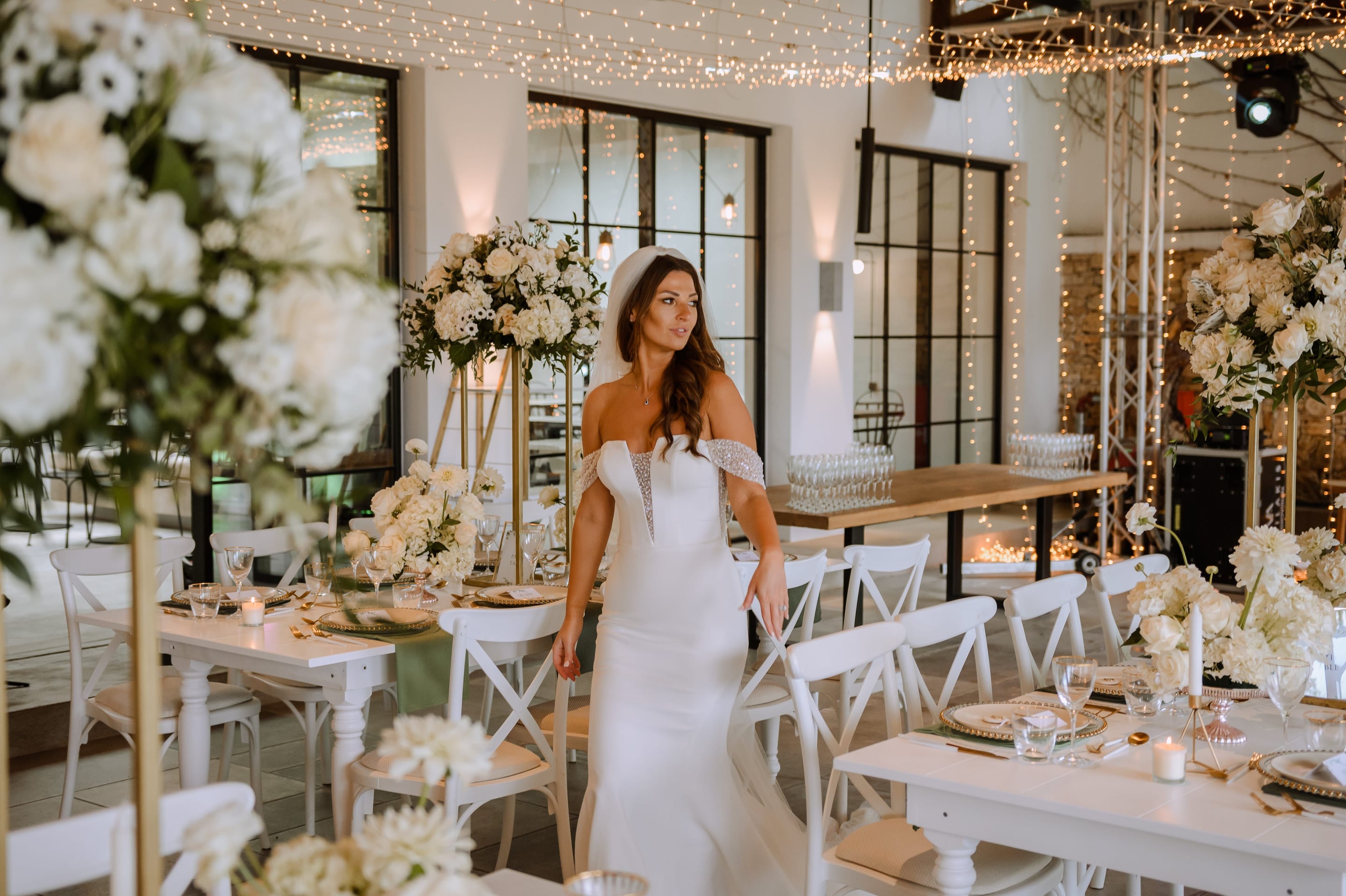 A bride in a fitted white satin gown with beaded off-shoulder straps stands among white cross-back chairs in a bright reception room, where tall gold stands hold lush white hydrangeas, anemones and roses. Strings of warm fairy lights hang overhead, and the scene is a wedding table setting at Minos Beach Art Hotel in Greece.