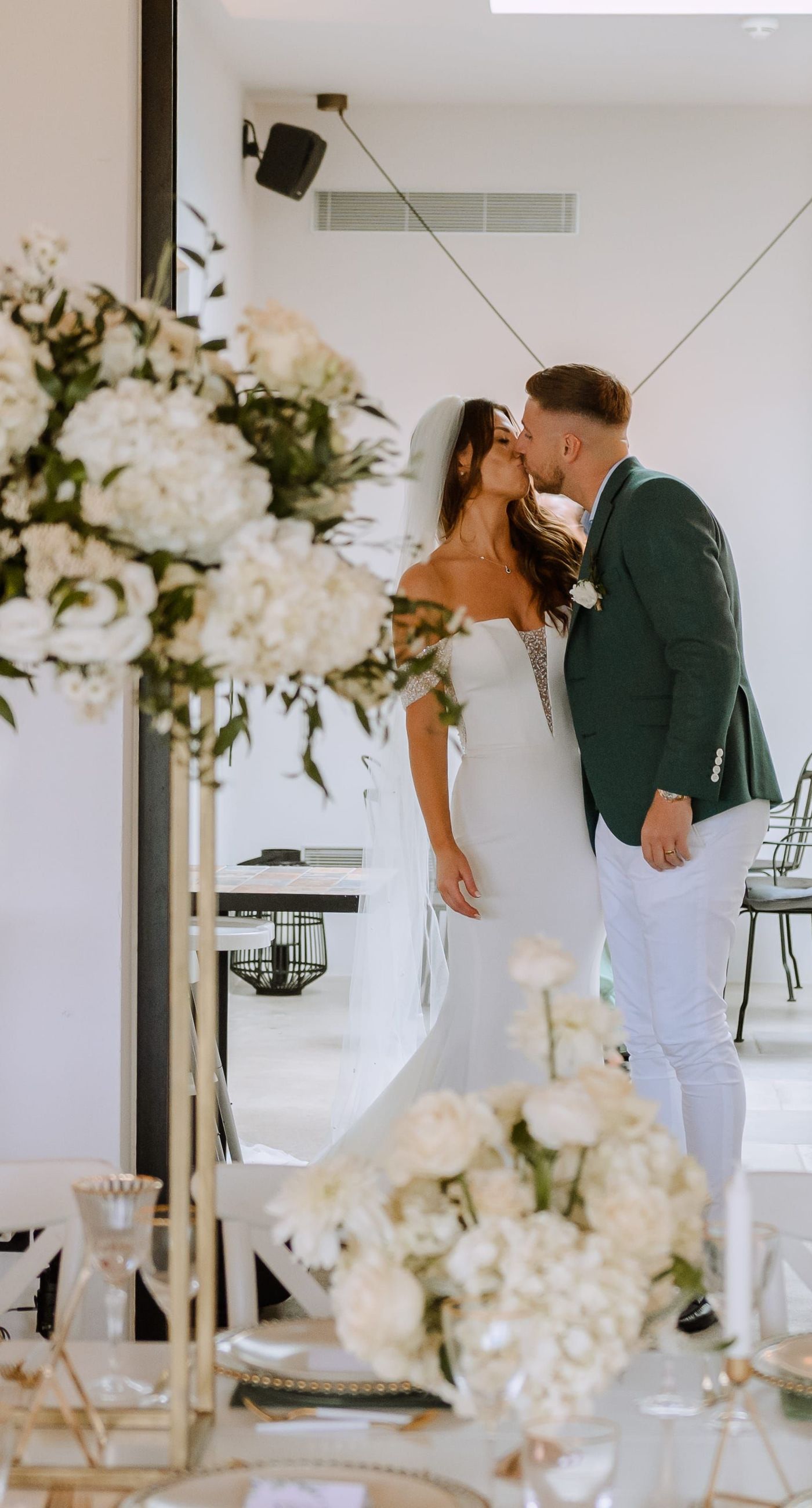 Christine & Eduart - Under a delicate floral arch, adorned with lush white blossoms and greenery, stands a vision of elegance and romance. The gentle pastel hues of the surrounding florals beautifully complement the soft, ethereal ambiance of this enchanting scene. It feels as though nature itself has been curated to frame a moment of serene connection, blending seamlessly into a verdant backdrop. The contrast of the bride's exquisite, flowing gown with the groom's tailored, modern attire in shades of emerald and white speaks volumes of both tradition and contemporary flair. This work captures an essence of timeless beauty, harmoniously blending natural elements with sophisticated design. - Photo 2