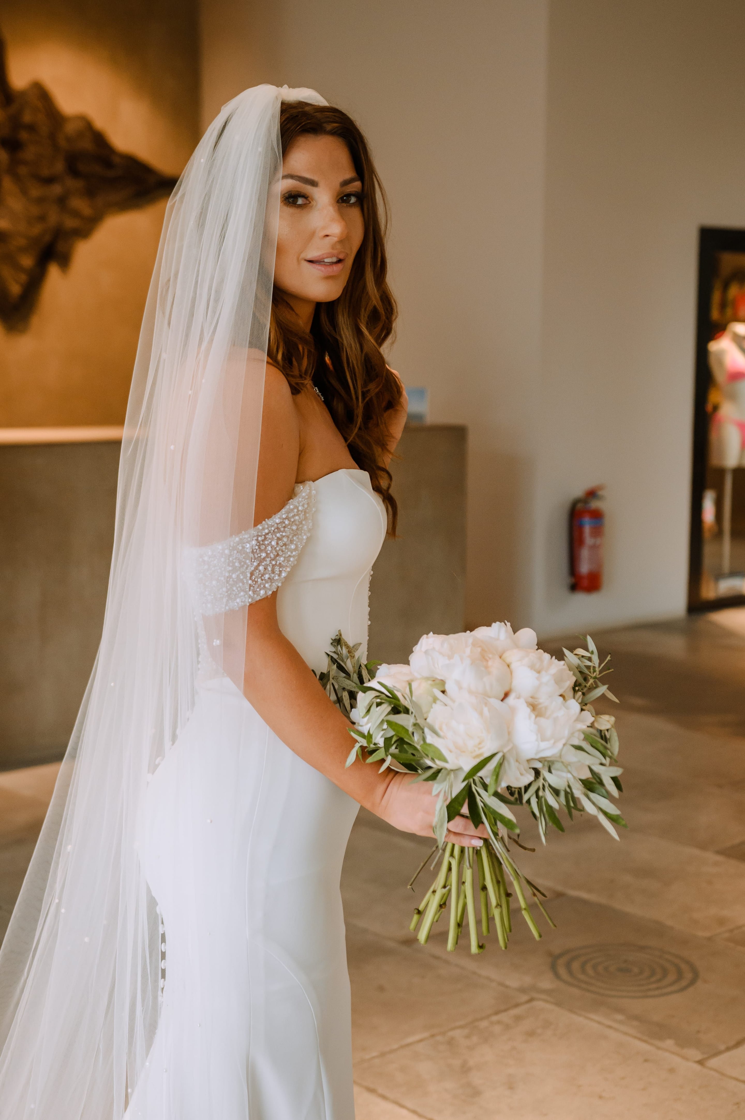 A bride with long, softly waved brown hair and a sheer veil glances back over her shoulder, holding a full bouquet of white peonies and olive sprigs. Her strapless satin gown has a beaded, off-the-shoulder detail, and she stands in a warm, light-filled hallway at Minos Beach Art Hotel in Greece. The wedding bouquet's cream and green tones echo the soft, neutral setting.