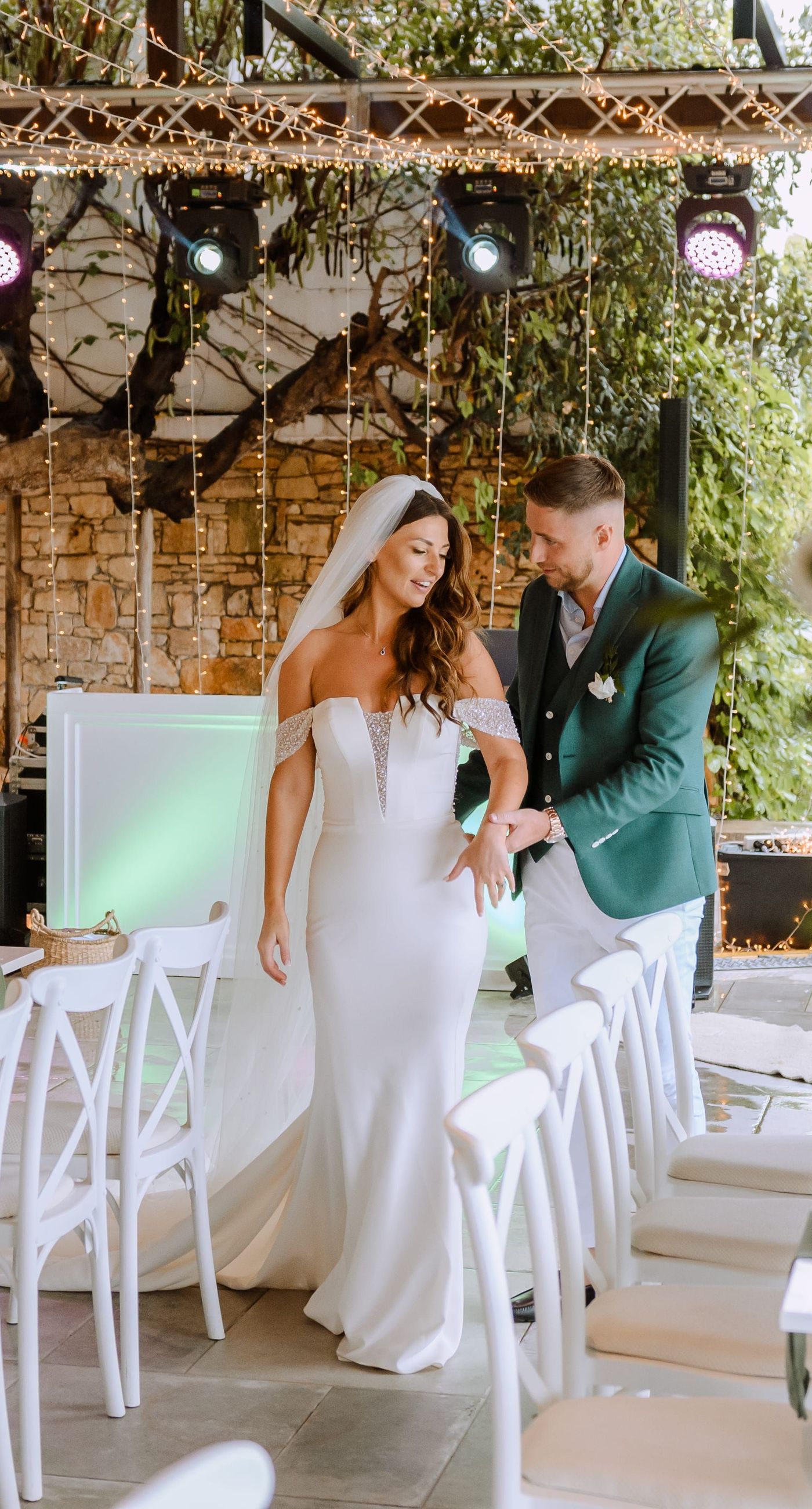 Christine & Eduart - Under a delicate floral arch, adorned with lush white blossoms and greenery, stands a vision of elegance and romance. The gentle pastel hues of the surrounding florals beautifully complement the soft, ethereal ambiance of this enchanting scene. It feels as though nature itself has been curated to frame a moment of serene connection, blending seamlessly into a verdant backdrop. The contrast of the bride's exquisite, flowing gown with the groom's tailored, modern attire in shades of emerald and white speaks volumes of both tradition and contemporary flair. This work captures an essence of timeless beauty, harmoniously blending natural elements with sophisticated design. - Photo 7