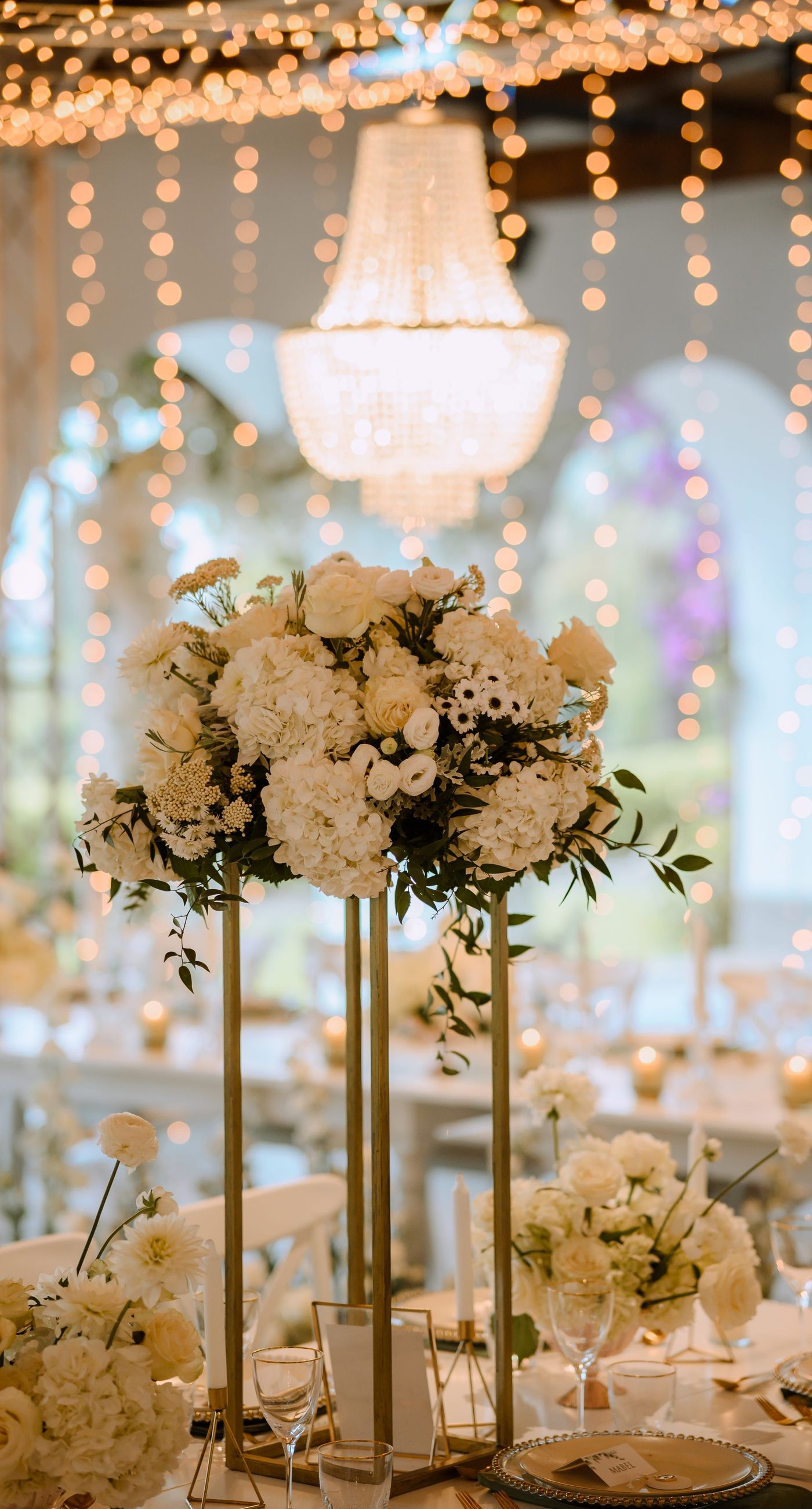 Christine & Eduart - Under a delicate floral arch, adorned with lush white blossoms and greenery, stands a vision of elegance and romance. The gentle pastel hues of the surrounding florals beautifully complement the soft, ethereal ambiance of this enchanting scene. It feels as though nature itself has been curated to frame a moment of serene connection, blending seamlessly into a verdant backdrop. The contrast of the bride's exquisite, flowing gown with the groom's tailored, modern attire in shades of emerald and white speaks volumes of both tradition and contemporary flair. This work captures an essence of timeless beauty, harmoniously blending natural elements with sophisticated design. - Photo 5