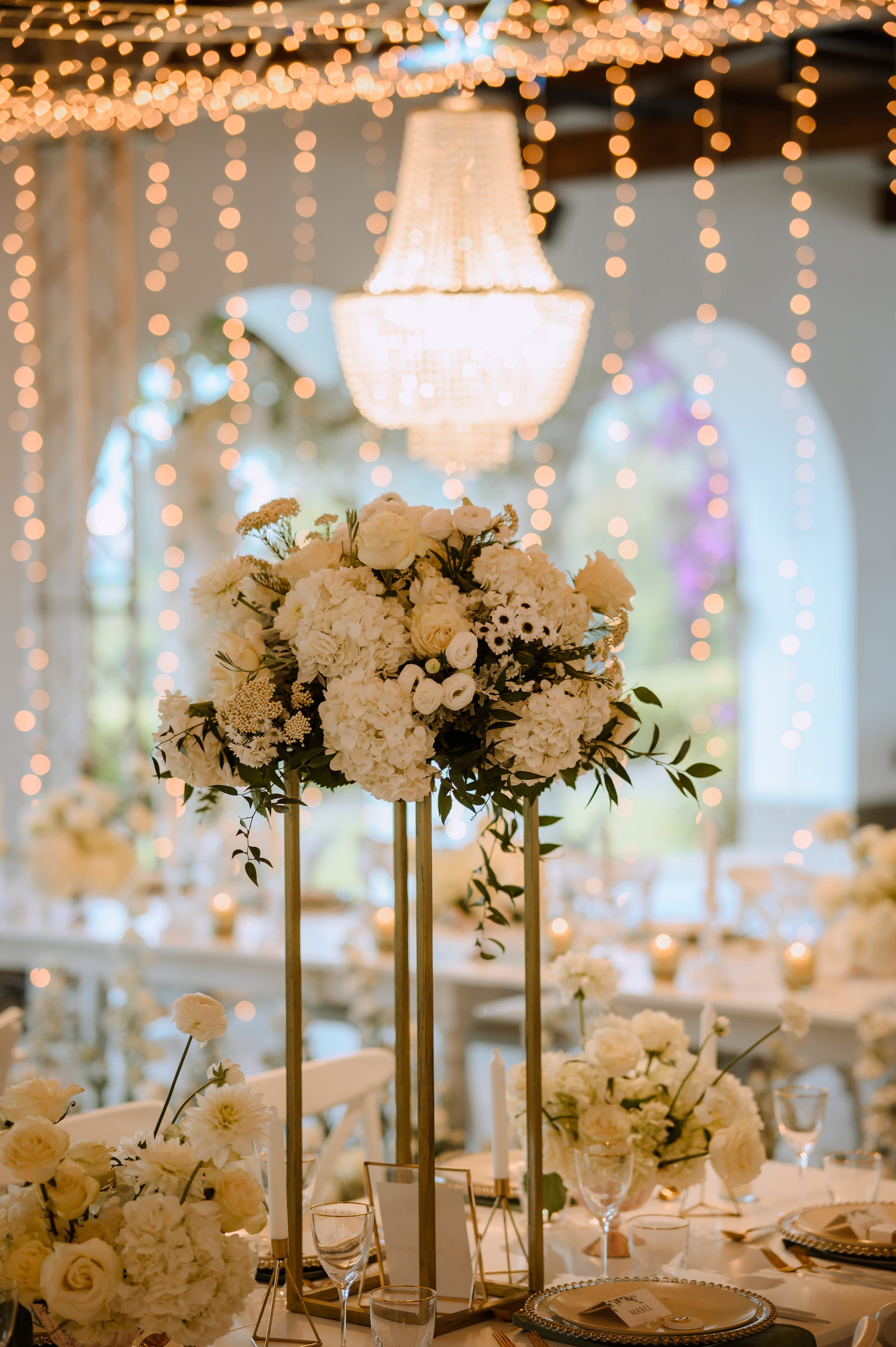 A tall wedding centrepiece of white hydrangeas, cream garden roses and small white blooms, with sprigs of green foliage trailing down, rests on slender gold stands at Minos Beach Art Hotel in Greece. Behind it, a crystal chandelier glows above strings of warm fairy lights, and soft candlelight fills the blurred arched windows and reception tables.