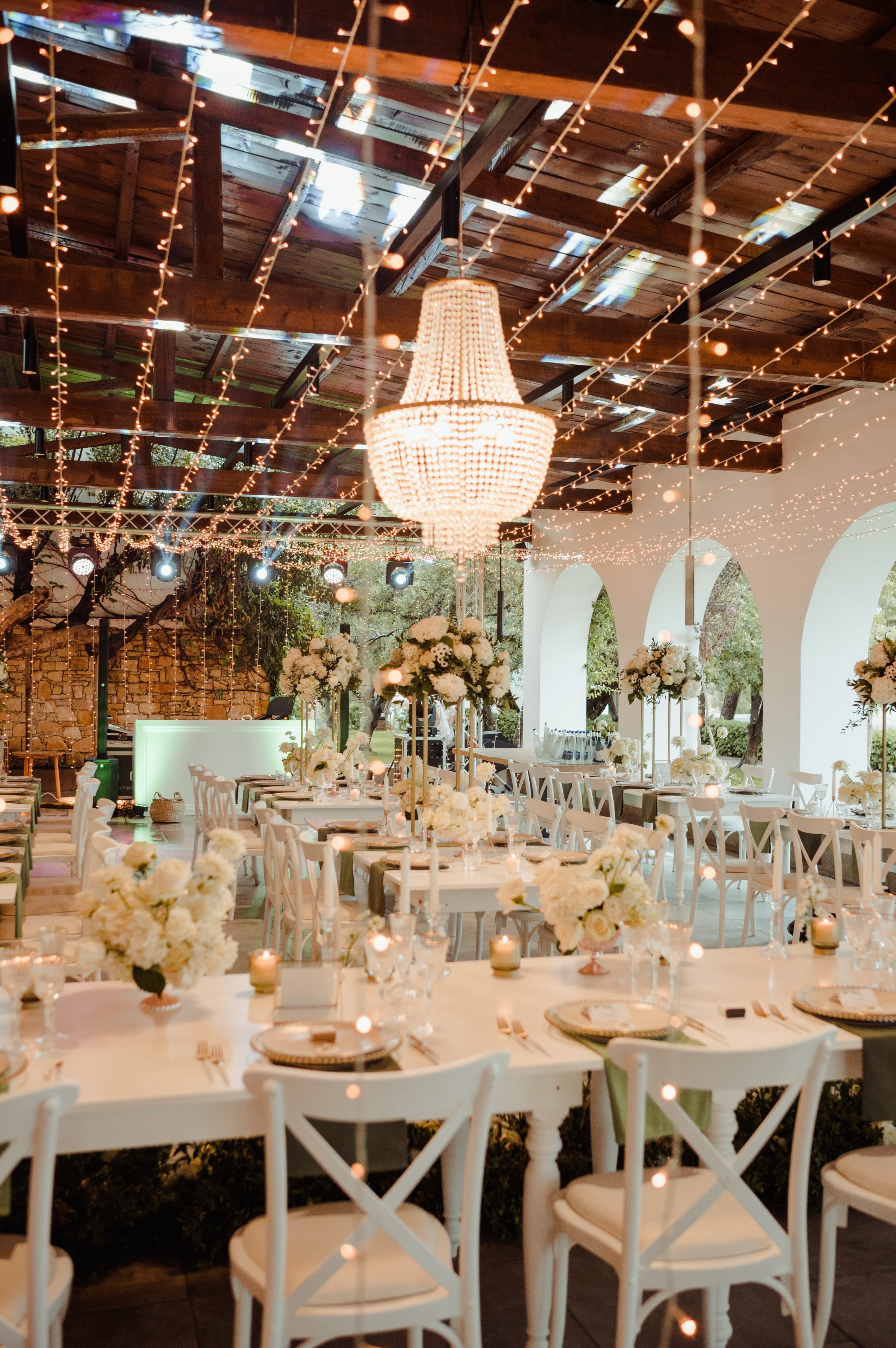 A wedding reception hall at Minos Beach Art Hotel in Greece, with a large beaded crystal chandelier hanging from a rustic timber ceiling strung with warm fairy lights. Long tables in white and sage linen are set with tall gold stands of white garden roses and hydrangeas, with more cream blooms along the floor, and white cross-back chairs line the room. Arched white walls open onto olive trees outside, giving the space a soft, candlelit, romantic glow.