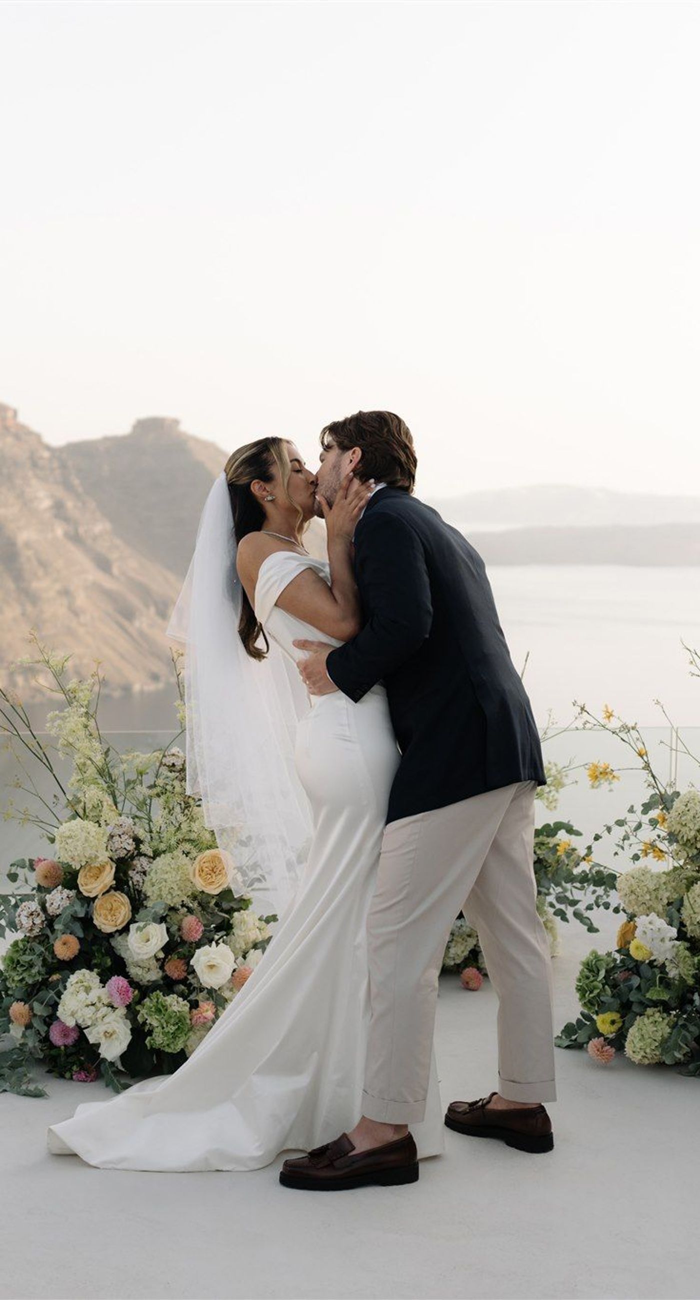 Courtney& Andrew - Bathed in the soft glow of a mountain backdrop, the scene captures a whimsical celebration of love. Soft white and blush rose petals float gently through the air, complementing the serene elegance of a bride in off-shoulder ivory silk and a groom in a classic black suit and tan trousers. The wooden chairs and delicate floral arrangements echo a natural, rustic charm, while the joyful guests add a vibrant energy. This portfolio piece beautifully encapsulates the essence of romantic bliss and harmonious design, set against nature's serene canvas. - Photo 8