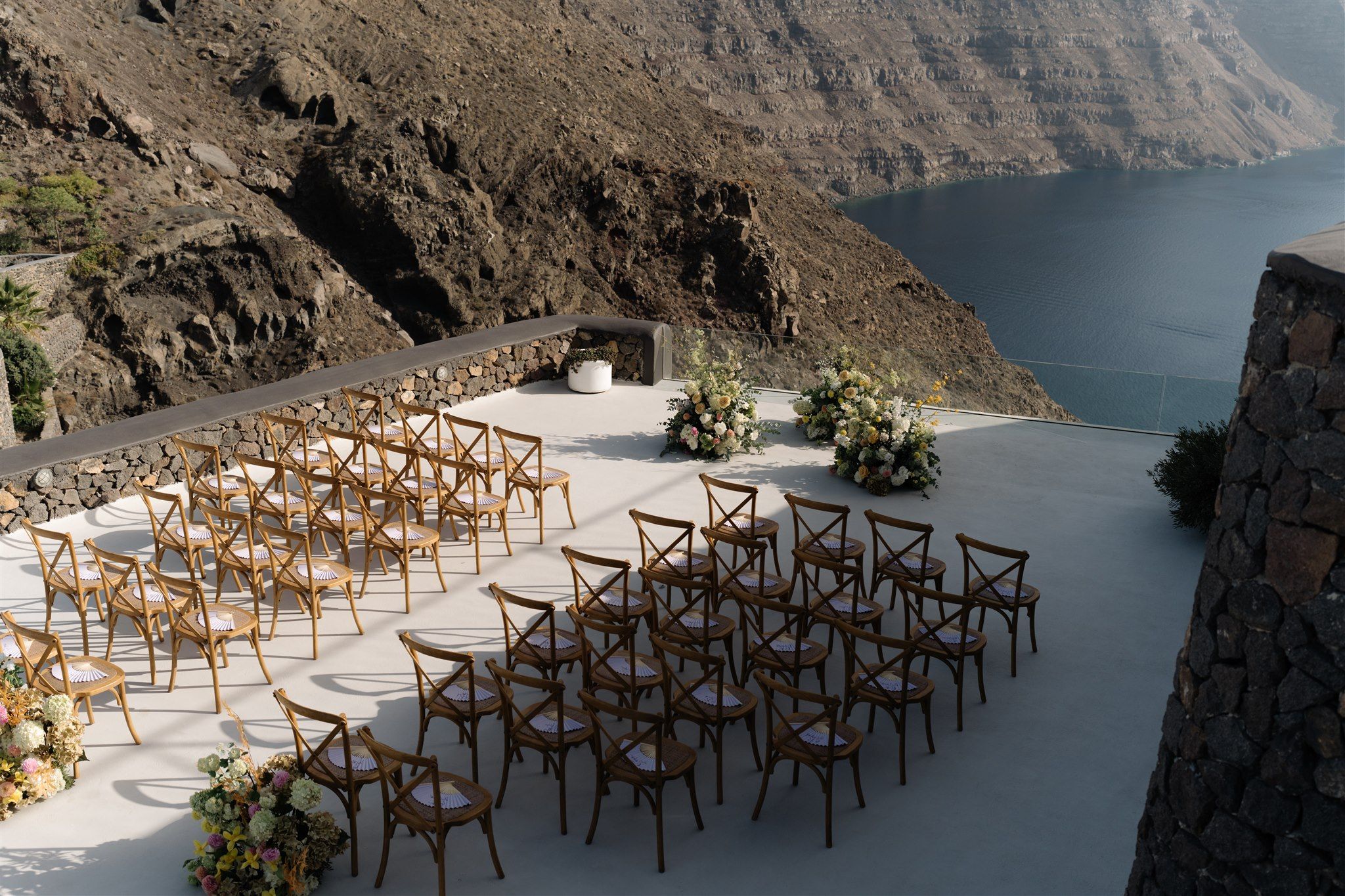 Rows of wooden cross-back chairs, each with a woven seat and a fan-folded white napkin, face a pale stone terrace set high on a volcanic cliff in Santorini, Greece, at Aenaon Villas. At the far edge, two lush arrangements of cream and blush blooms, with hydrangeas and soft yellow flowers, flank a low white urn, while the deep blue sea stretches beyond the rugged brown rock. These are wedding ceremony flowers for Courtney and Andrew's celebration.