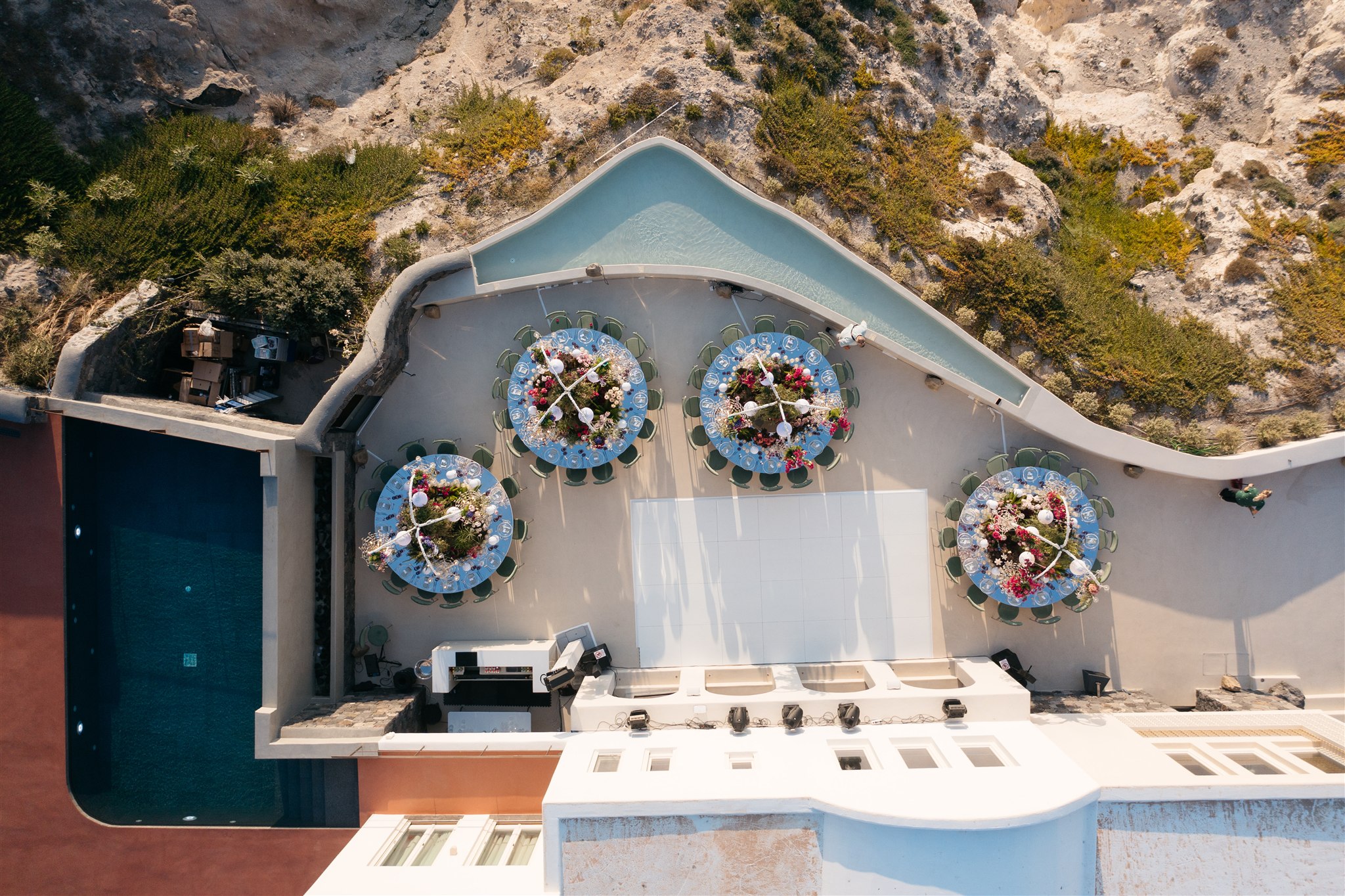 Seen from directly above, four round tables dressed in pale blue sit on a whitewashed terrace at iIslet in Santorini, Greece, each ringed with green chairs and crowned with a wild mass of pink, red and cream flowers and white orbs. Wedding table flowers like these spill over the centres while a pool glows turquoise along the edge, with rocky hillside scrub and olive-green shrubs rising behind. The midday light is bright and clear, giving the scene a calm, sun-washed feel.