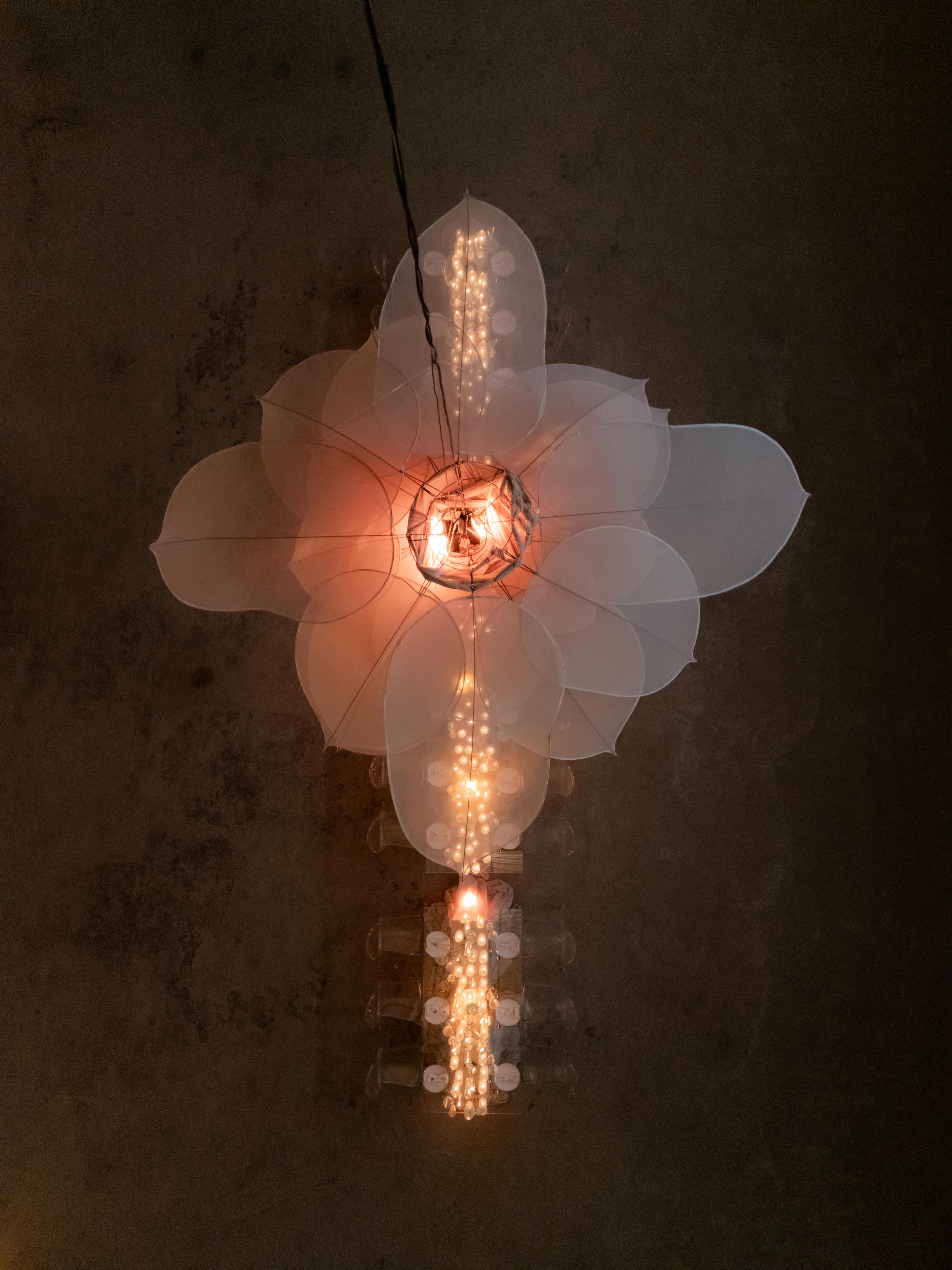 Overhead view of a large suspended flower installation of translucent, cream-white petals layered like an open bloom, glowing warmly from a lit core at its centre. Beneath it, strings of small glowing beads and clusters of lights hang down over a long table set with glasses and candles, all against a dark stone wall. This is an event floral and lighting installation in Rethimno, Crete, Greece.