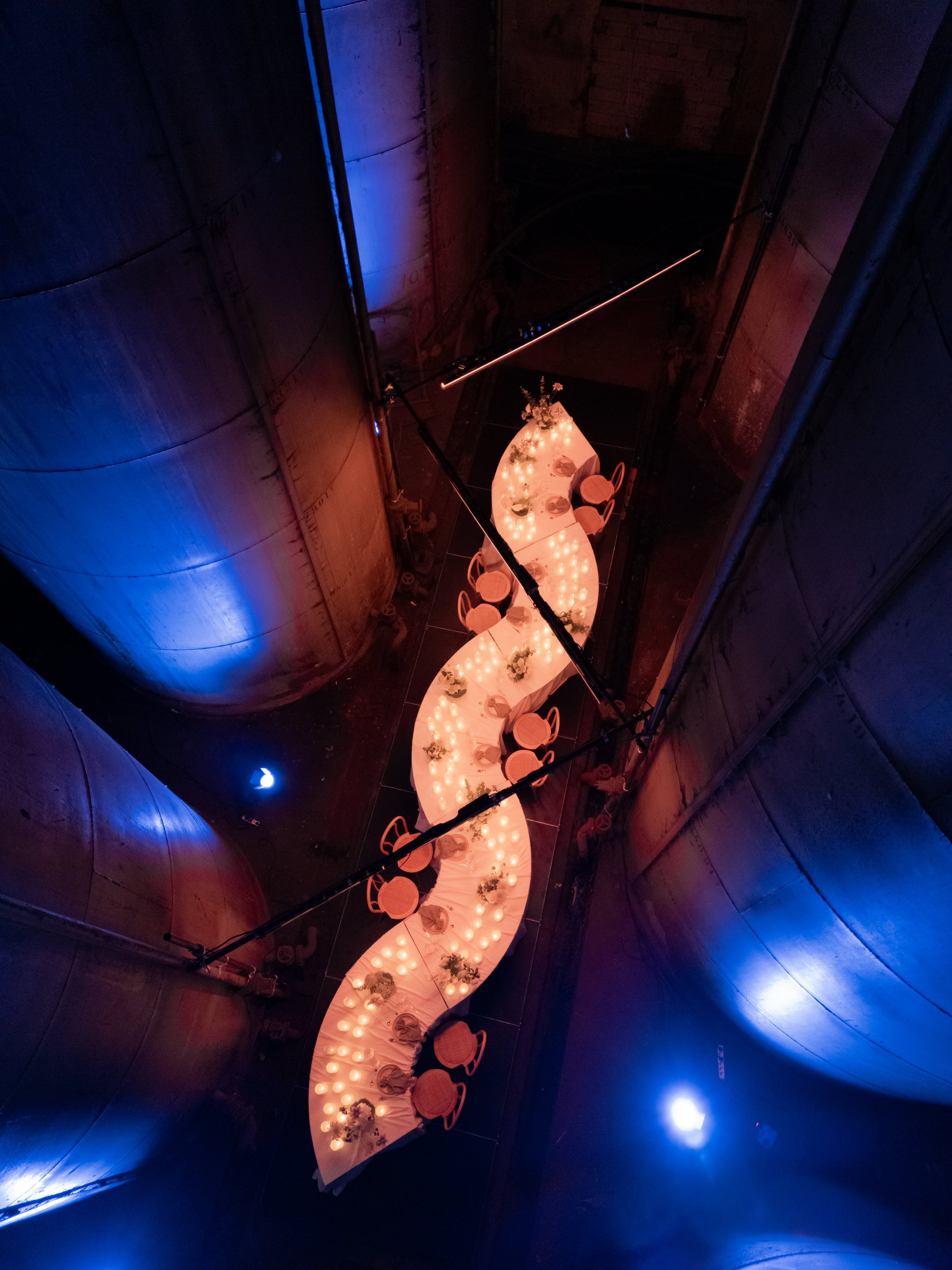 A long, S-shaped banquet table draped in white cloth runs diagonally through a vast industrial space, its edges lined with rows of glowing candles and small floral centrepieces of white and green foliage. Terracotta-coloured chairs sit along both sides, and the photo is taken from above, so the table seems to snake between curved walls lit in deep blue and warm amber. This is a wedding table setting in Rethimno, Crete, Greece.