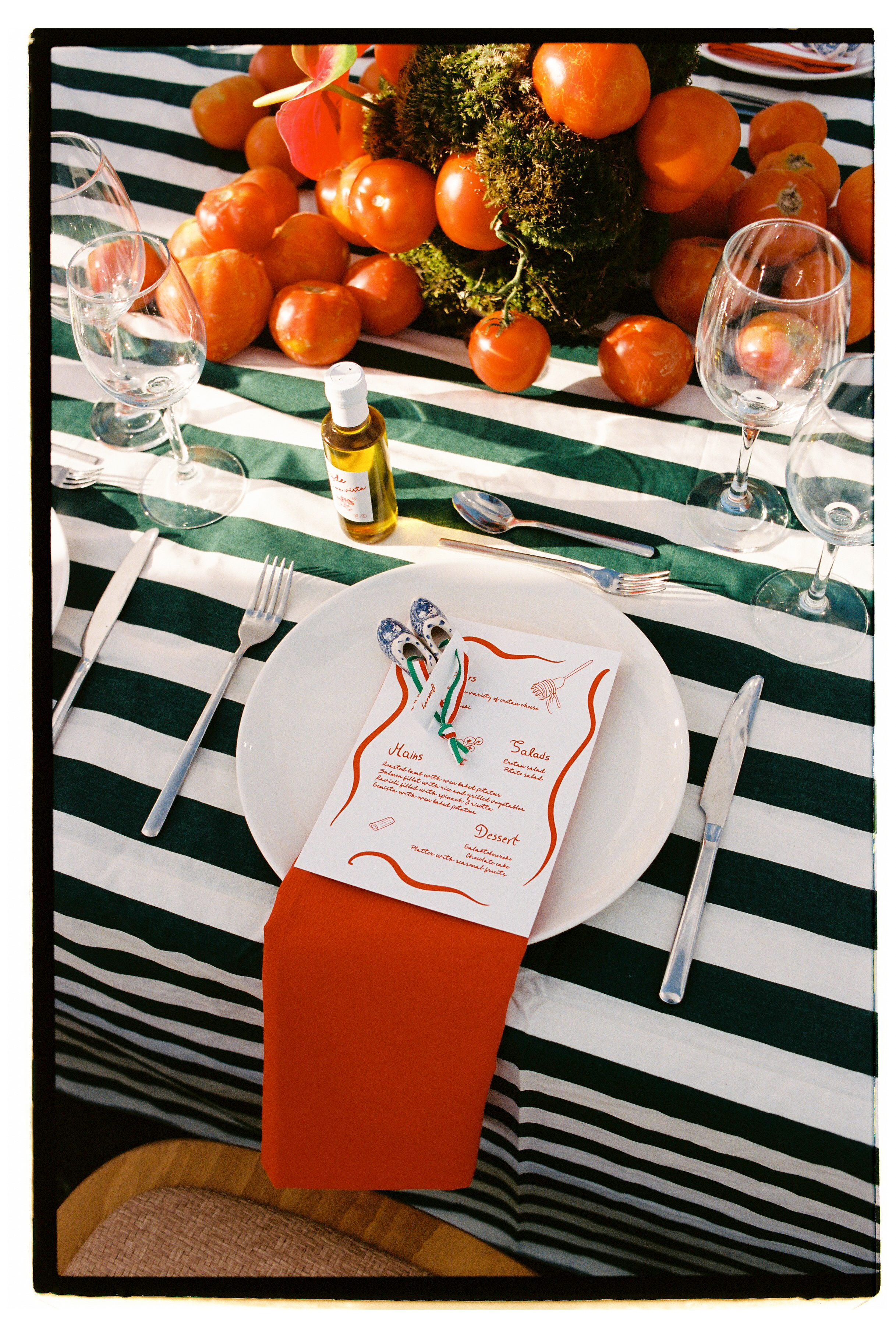 A wedding table in Greece, set at Wine Dark Villas, with a green and white striped runner dotted with ripe tomatoes and moss-wrapped centrepiece. A white plate sits on an orange napkin beneath a hand-lettered menu with wavy orange borders, tied with a green and white ribbon and two small blue and white ceramic shoes. Clear wine glasses, silver cutlery and a small bottle of olive oil complete the sunlit, rustic scene.