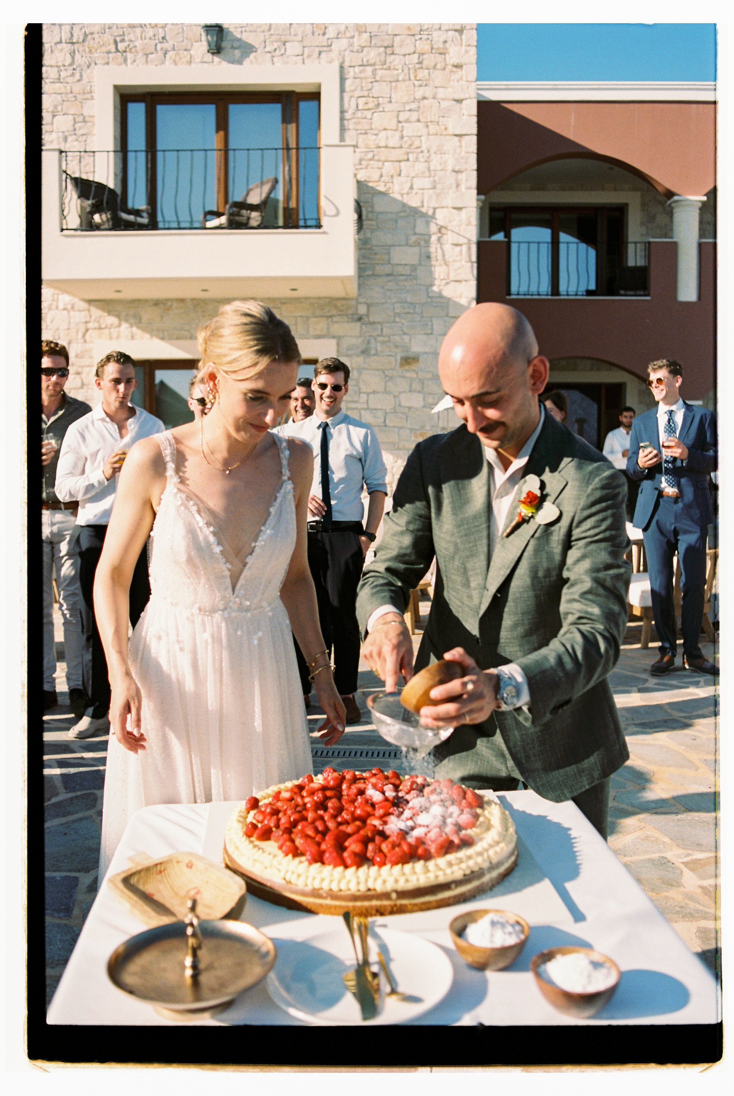 At a wedding reception at Wine Dark Villas in Greece, a bride in a pale, sleeveless gown with a deep V and delicate beaded detail stands beside a groom in a grey-green linen suit, who is dusting a strawberry tart with powdered sugar through a small sieve. The round tart is topped with rows of ripe red strawberries and sits on a white-draped table with small wooden bowls of sugar and a brass cake stand. Guests in smart summer attire gather behind them on a sunlit stone terrace.
