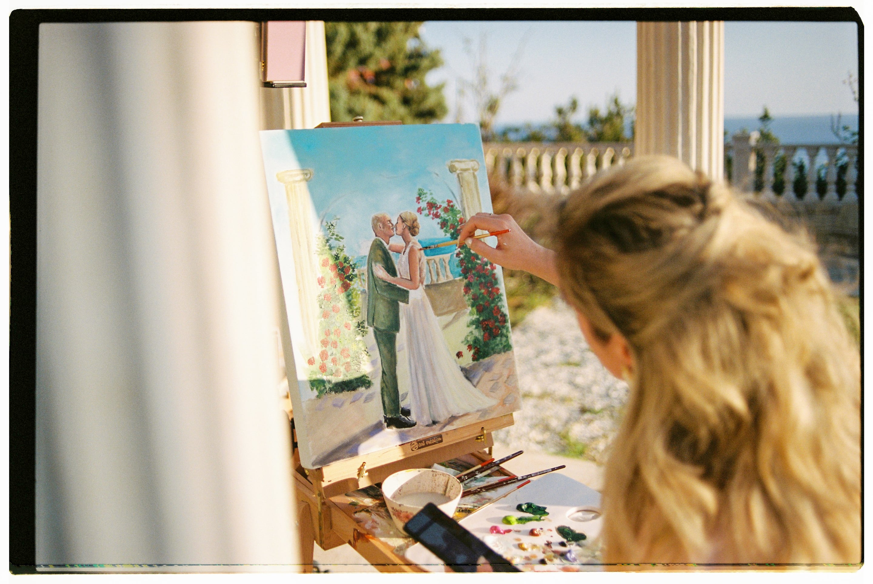 A painter with her blonde hair pulled back works at an easel outdoors, brush raised to a canvas showing a bride in a flowing white gown kissing a groom in a green suit beside a rose-covered arch and classical columns, with a blue sea behind them. Palettes of paint and a bowl of water sit on the table, and the warm sunlight suggests a wedding painting being made live at Wine Dark Villas in Greece for the Garden Ceremony story.