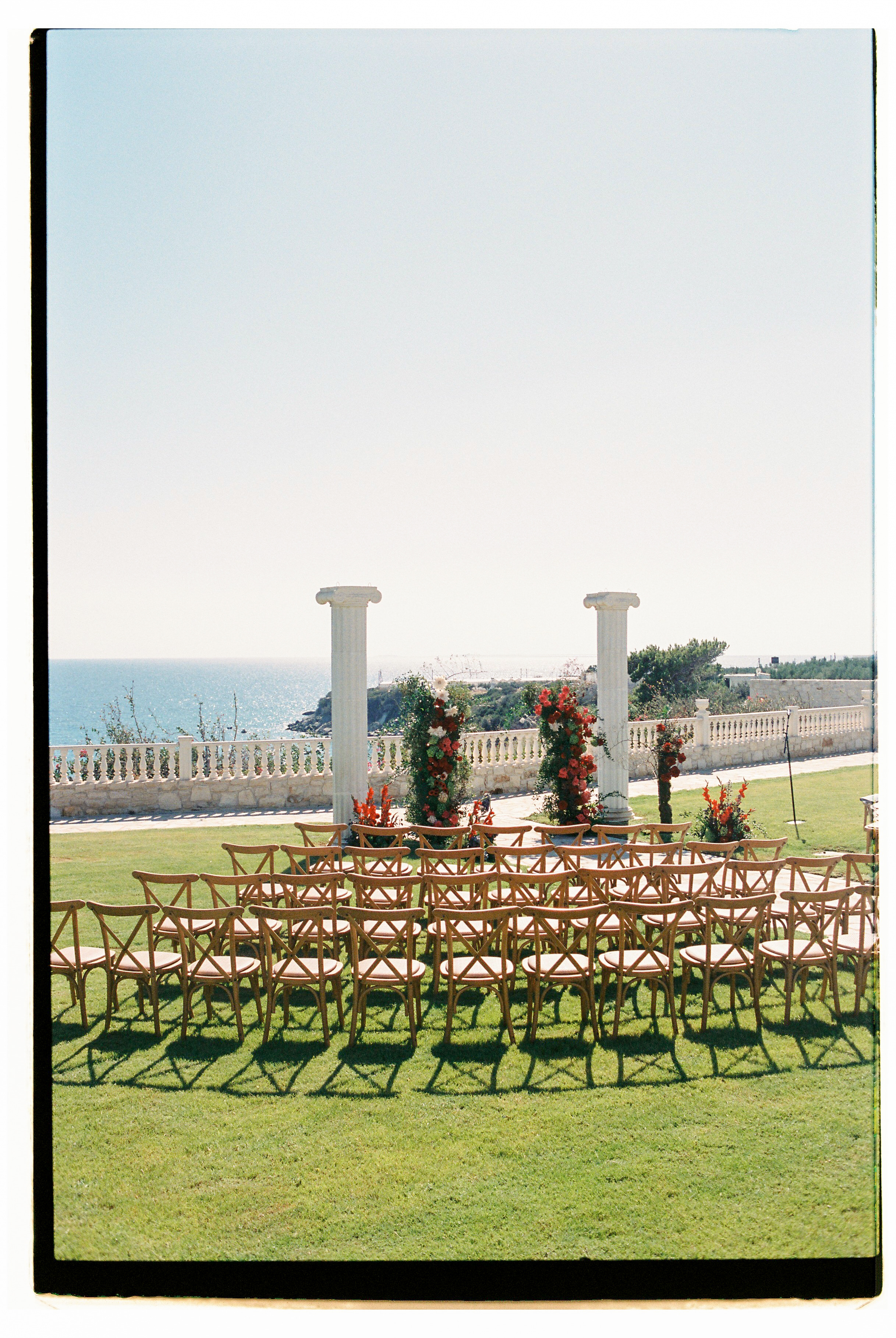 Rows of wooden cross-back chairs with pale cushions stand on a lush green lawn, casting long shadows in the sun, facing a ceremony arch dressed in deep red blooms and greenery, with orange gladioli at its base. Two tall white columns frame the scene against a stone balustrade and the blue sea beyond. This is a wedding ceremony set-up at Wine Dark Villas in Greece, with the flowers arranged for the aisle and altar.