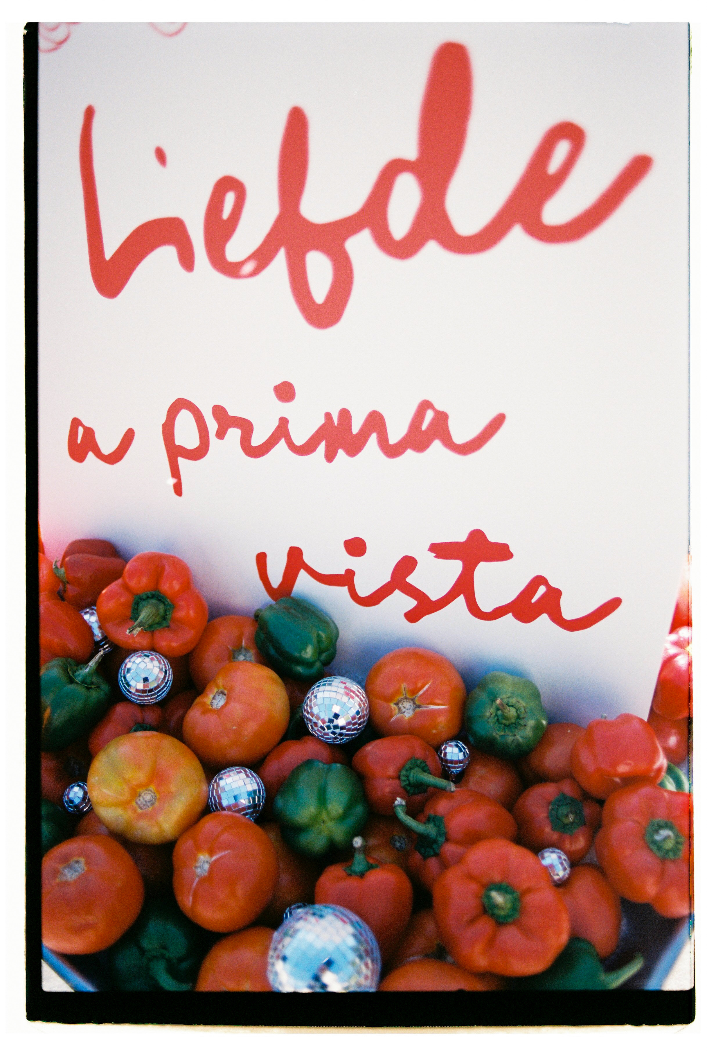 Red tomatoes and red and green peppers are heaped in a crate, scattered among small silver mirror disco balls that catch the light. Red hand-lettered script on the white sign behind them reads "vista," with a partial word cut off above it. The film-grain close-up has a playful, retro mood, and it is a styling image for Wine Dark Villas in Greece rather than a floral arrangement.