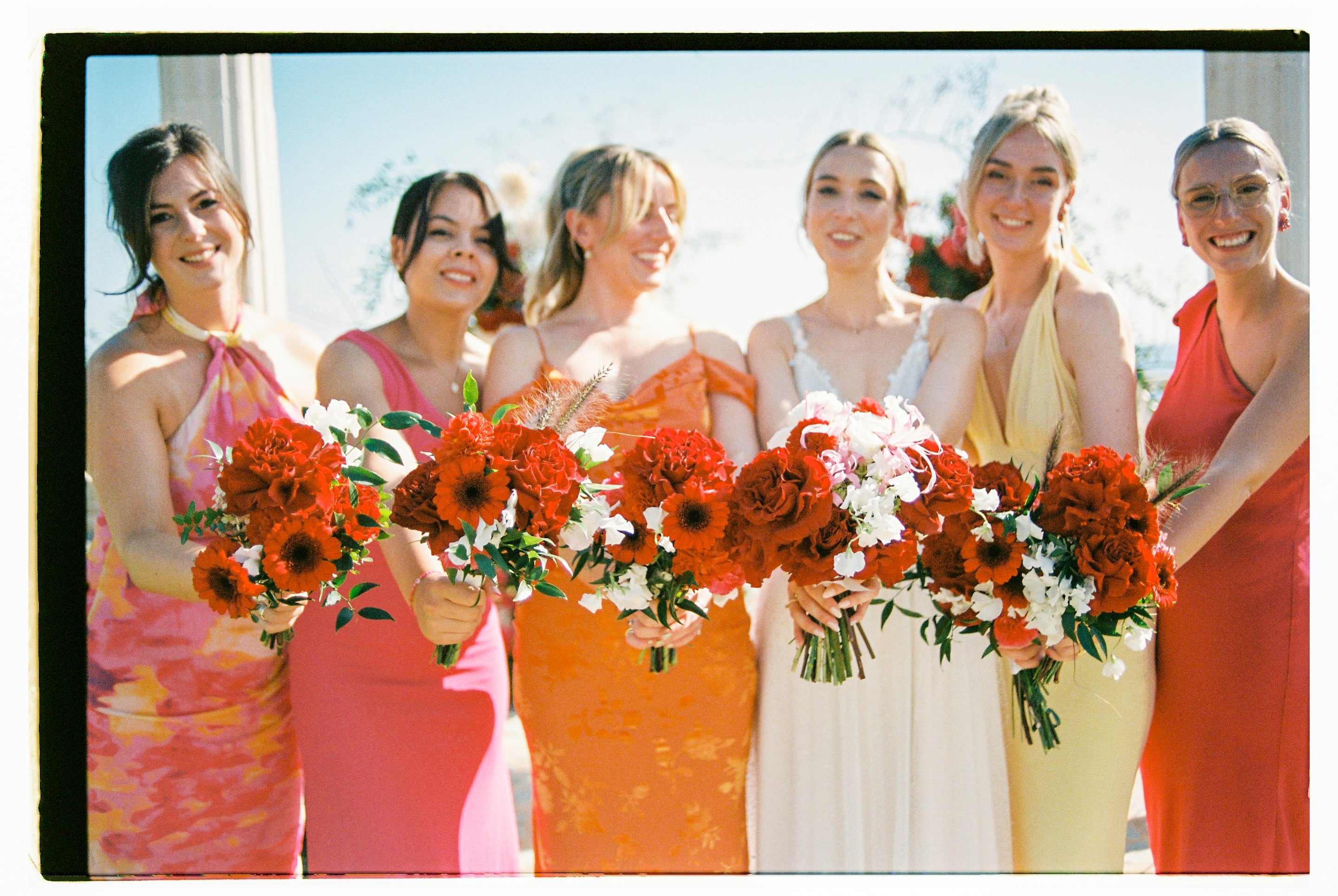 Five smiling bridesmaids in dresses shading from pink to orange to pale yellow hold out bouquets of deep red garden roses, red gerbera daisies and carnations, tucked with white sweet peas and small green leaves, at a wedding flower arrangement at Wine Dark Villas in Greece. The warm film-like tones and bright blue sky make the scene feel sunny and relaxed.