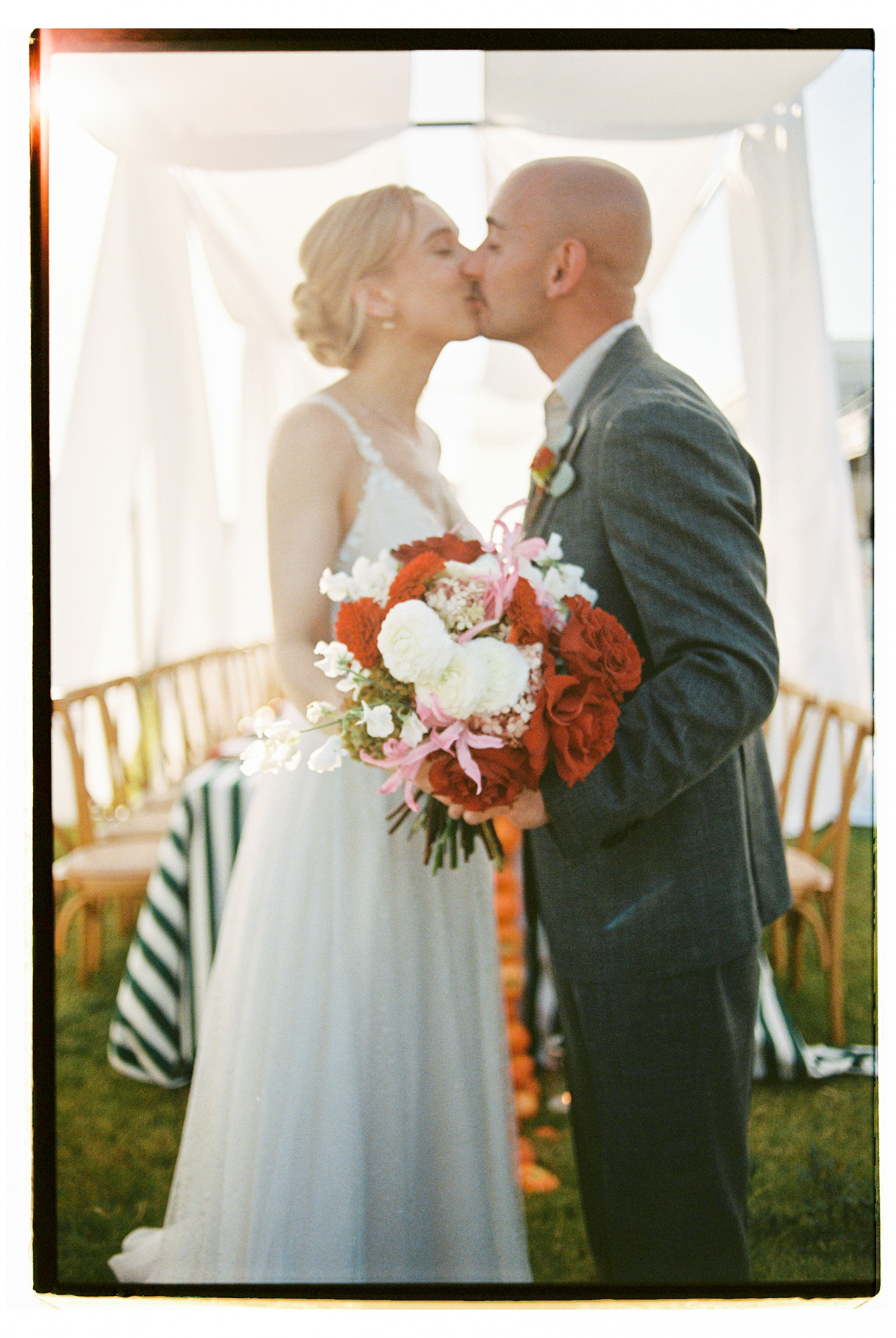 A bridal bouquet of deep red garden roses, white ranunculus, pink lily blooms and sweet peas, held by a bride in a pale tulle gown as she and her groom kiss behind it. The softly blurred background shows sheer white drapes and wooden chairs at a wedding ceremony in Greece, at Wine Dark Villas.