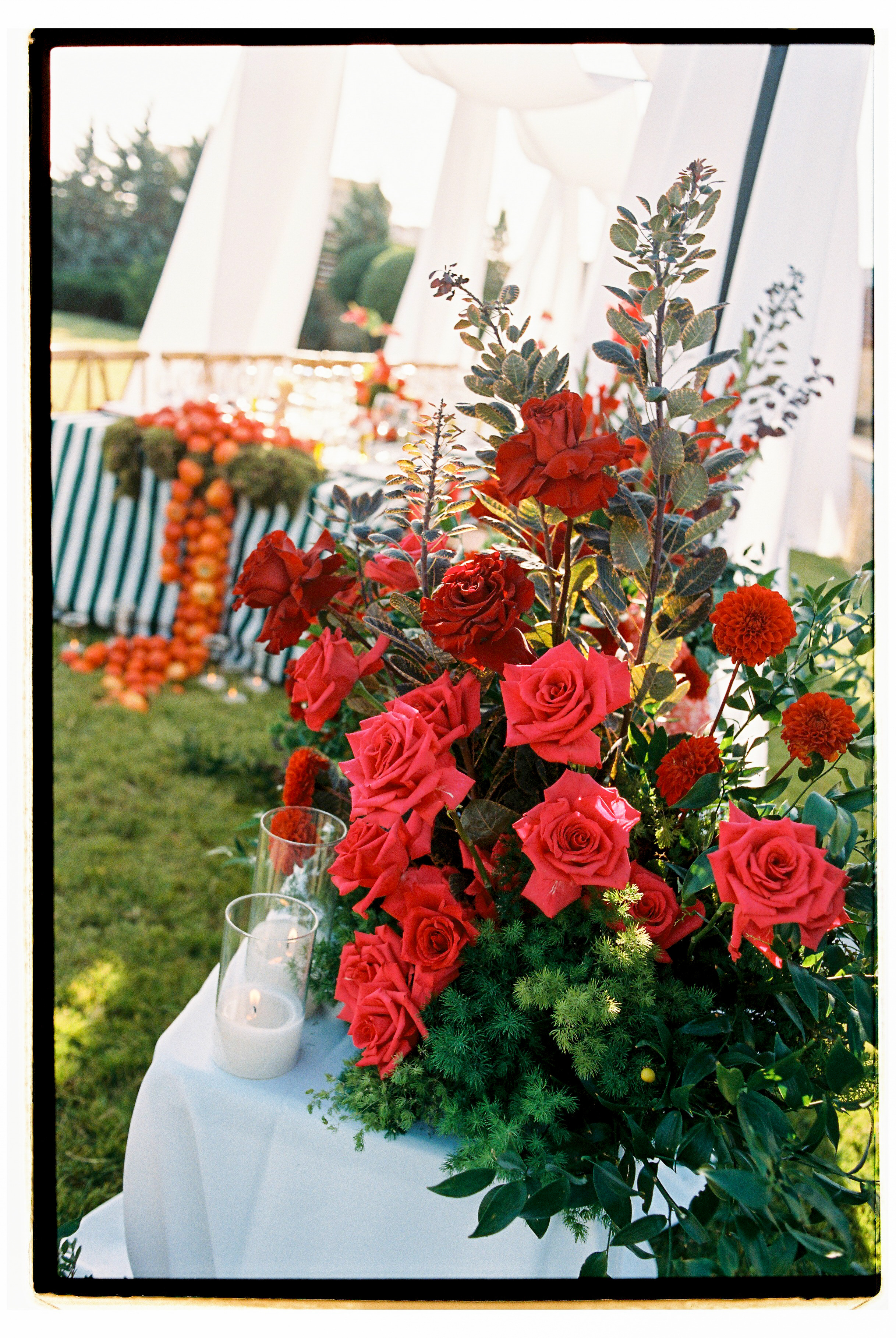 A tall, lush arrangement of red garden roses in shades from deep crimson to bright coral, mixed with burnt-orange dahlias and dark, bronze-tinted foliage, rising from a bed of feathery green sprigs. Beside it on a white-draped table stand two glass hurricane candles with lit tapers, and behind, a striped green-and-white tablecloth is lined with orange blooms beneath soft, out-of-focus light. This is a wedding garden ceremony at Wine Dark Villas in Greece, with the warm, romantic palette set against green lawn.