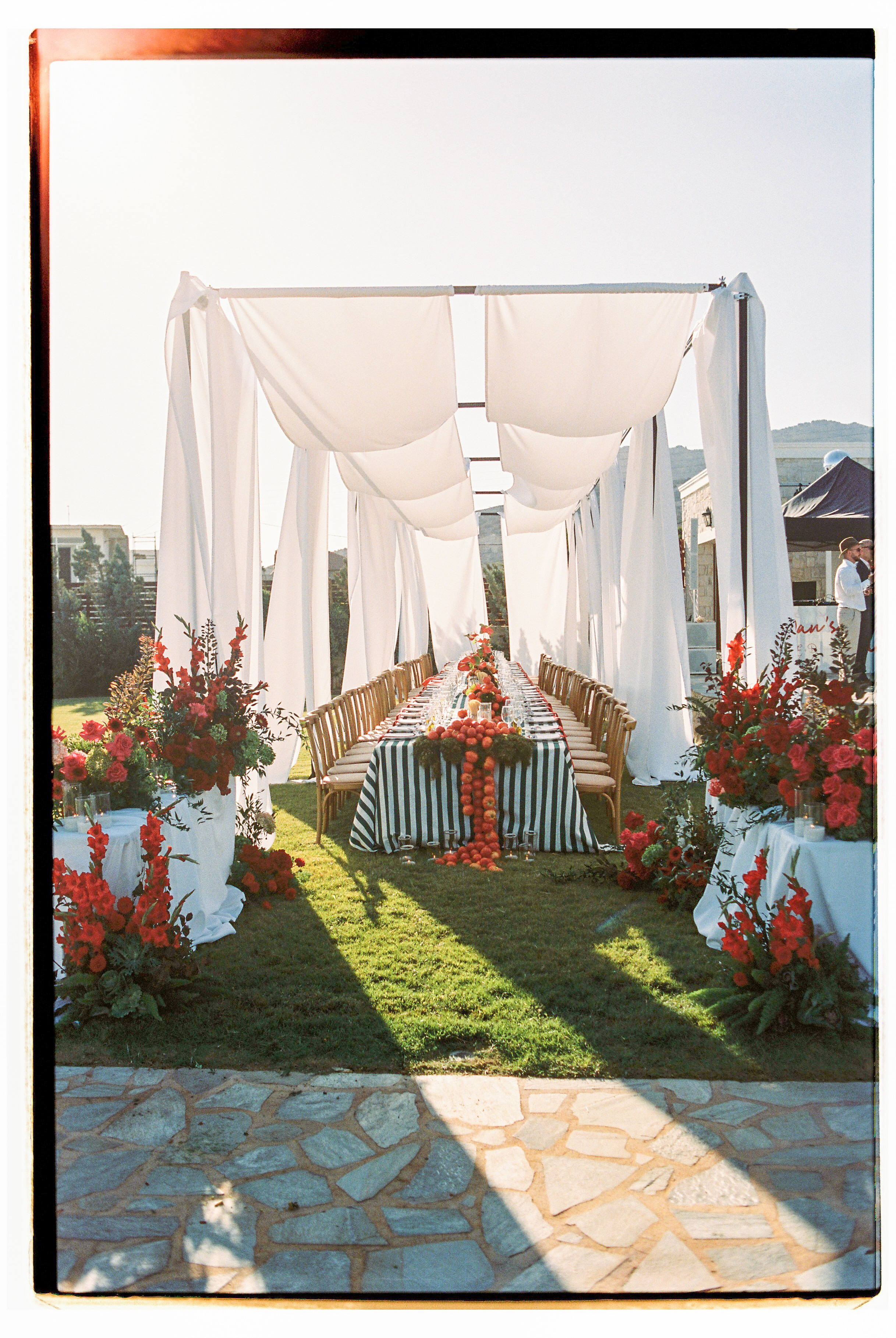 A long outdoor wedding table runs beneath a canopy of flowing white fabric, set on a lawn at Wine Dark Villas in Greece. Its green-and-white striped cloth is decorated with a runner of red and orange blooms and small apples, with tall red gladioli and rose arrangements standing at either end. Wooden chairs line both sides under soft, golden afternoon light.