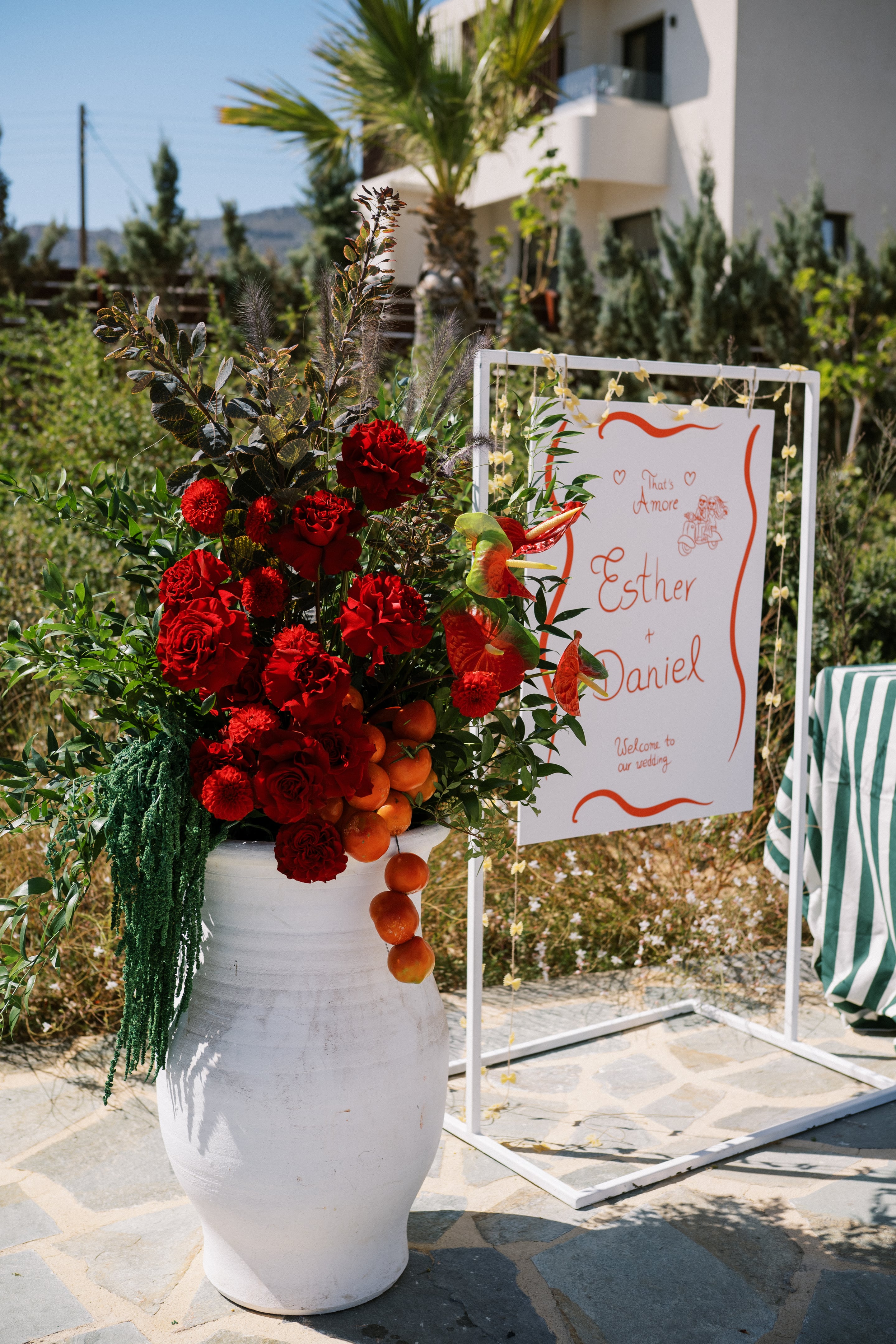 A tall white urn overflowing with deep red garden roses, dahlia-like red blooms, and red anthuriums, set among dark green foliage, grey plumes and small orange fruits at its base, all for a wedding ceremony at Wine Dark Villas in Greece. Beside it stands a white easel sign with red hand-lettered script reading "That's Amore, Esther + Daniel, Welcome to our wedding," framed by red wavy lines, with a garden of cypress trees and a white building behind under bright daylight.