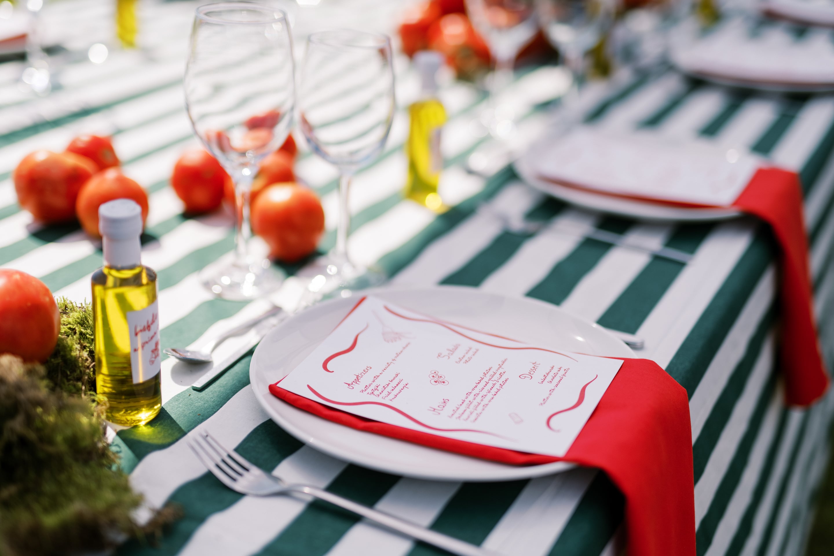 A place setting on a green-and-white striped tablecloth at Wine Dark Villas in Greece, styled for a wedding meal outdoors. A white plate sits under a cream menu card with handwritten red script and a red napkin folded beneath it, with clear wine glasses, small bottles of olive oil, and ripe red tomatoes scattered along the table. The bright daylight and soft blur of the background give it a warm, relaxed Mediterranean feel.