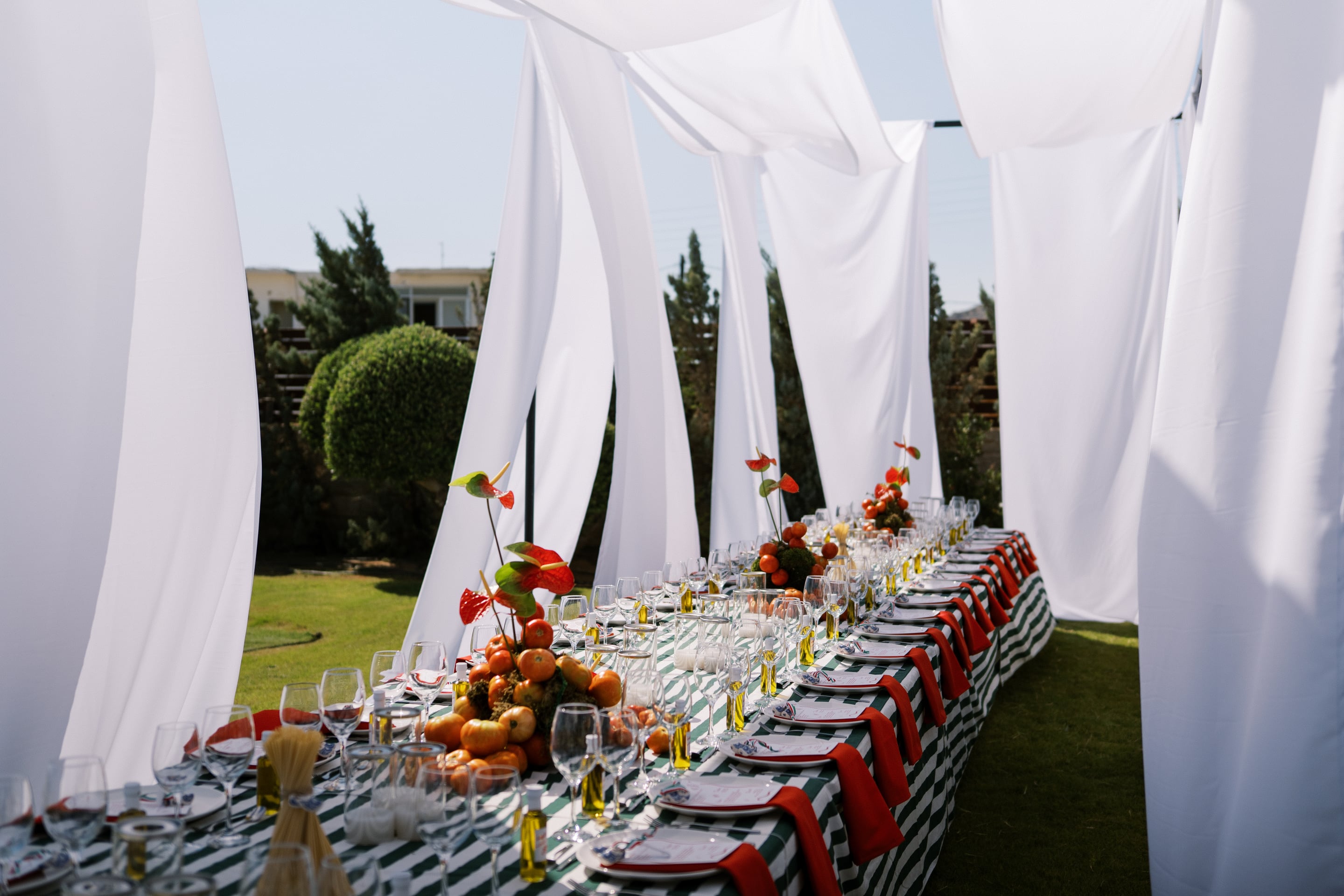 A long wedding banquet table in a garden at Wine Dark Villas in Greece, dressed in a green and white striped cloth with red napkins and rows of wine and water glasses. Clusters of ripe tomatoes and red anthurium flowers make the centrepieces, and billowing white fabric drapes overhead, filtering the afternoon sunlight. Trimmed green shrubs and trees sit behind, giving the scene a relaxed, sunny Mediterranean feel.