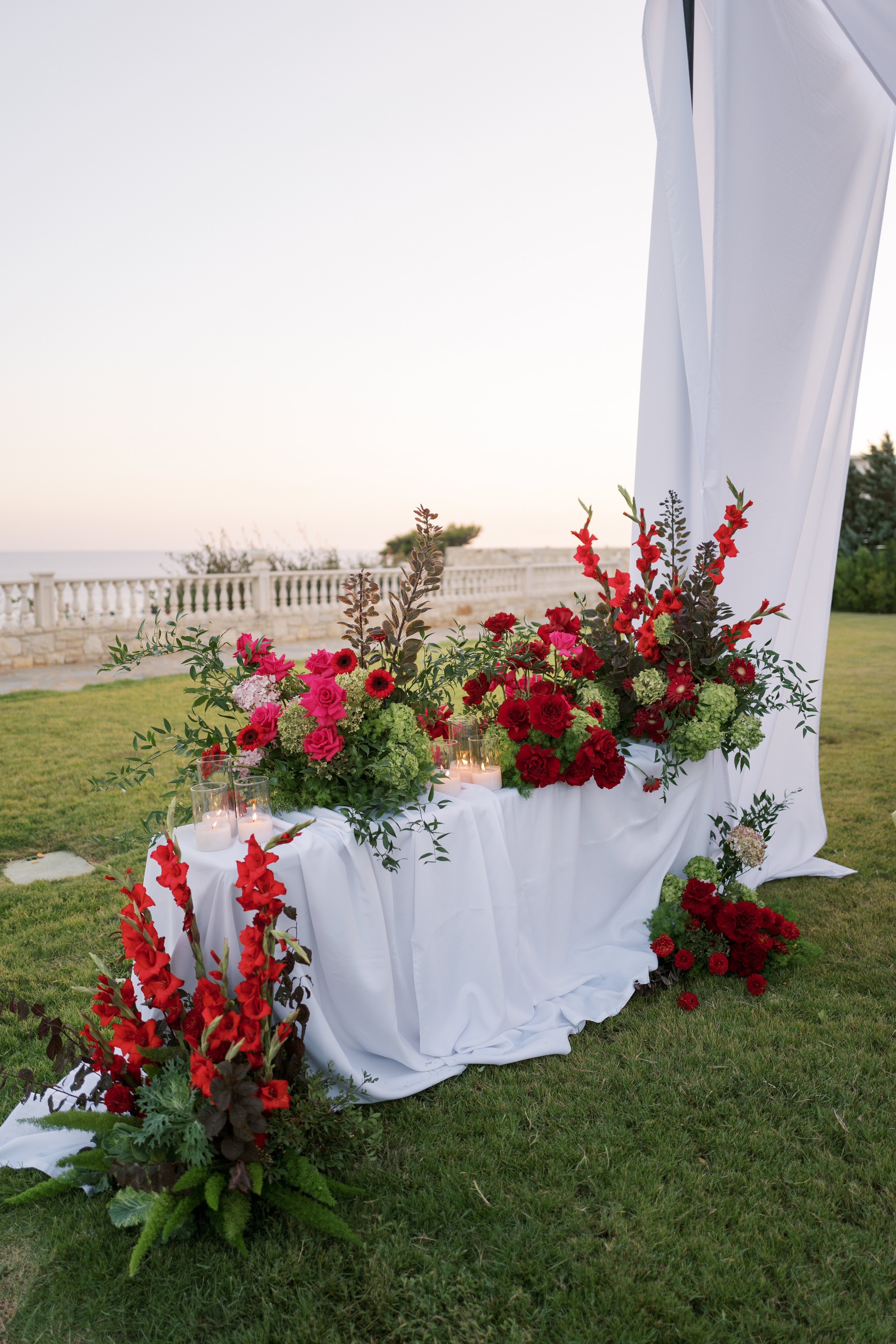 A long white draped table on a lawn at Wine Dark Villas in Greece, dressed with wedding flowers in red and hot pink: tall red gladioli, deep red roses and carnations, pink roses, and green hydrangeas, all set among dark foliage and trailing greenery. Clustered glass votive candles glow at one end, and behind the table a white fabric panel falls from a ceremony arch. Beyond a white balustrade, the sea and a soft pale sky fill the background in the early evening light.