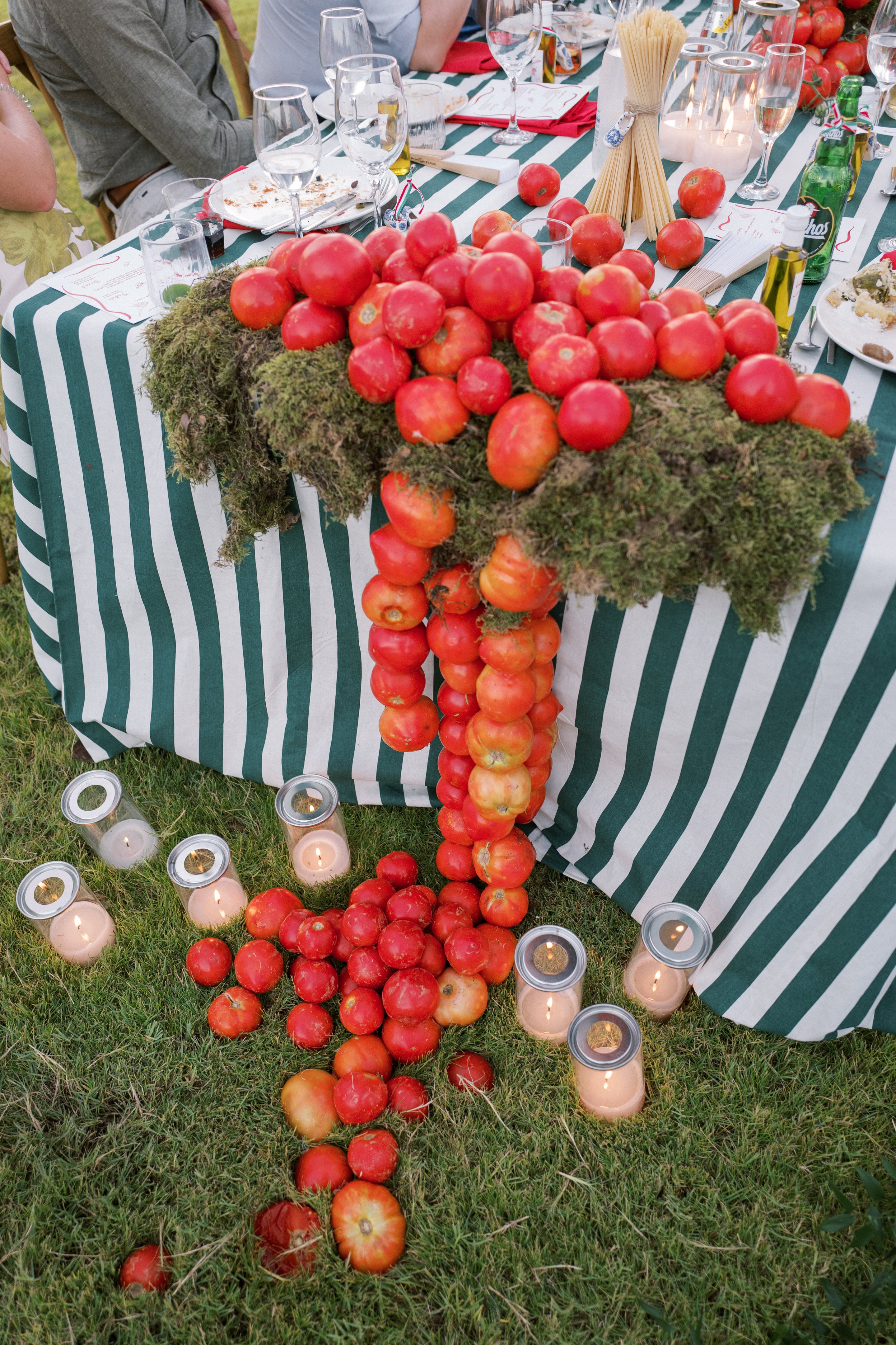 Cascades of ripe red tomatoes spill over a moss-covered green and white striped table at Wine Dark Villas in Greece, a wedding table display where the tomatoes tumble down the front in long strands. Scattered on the grass below are more tomatoes and several small glass hurricane lanterns, their candles glowing warmly in the evening light. The setting feels rustic and garden-like, with the bright red fruit standing out against the green moss and striped cloth.