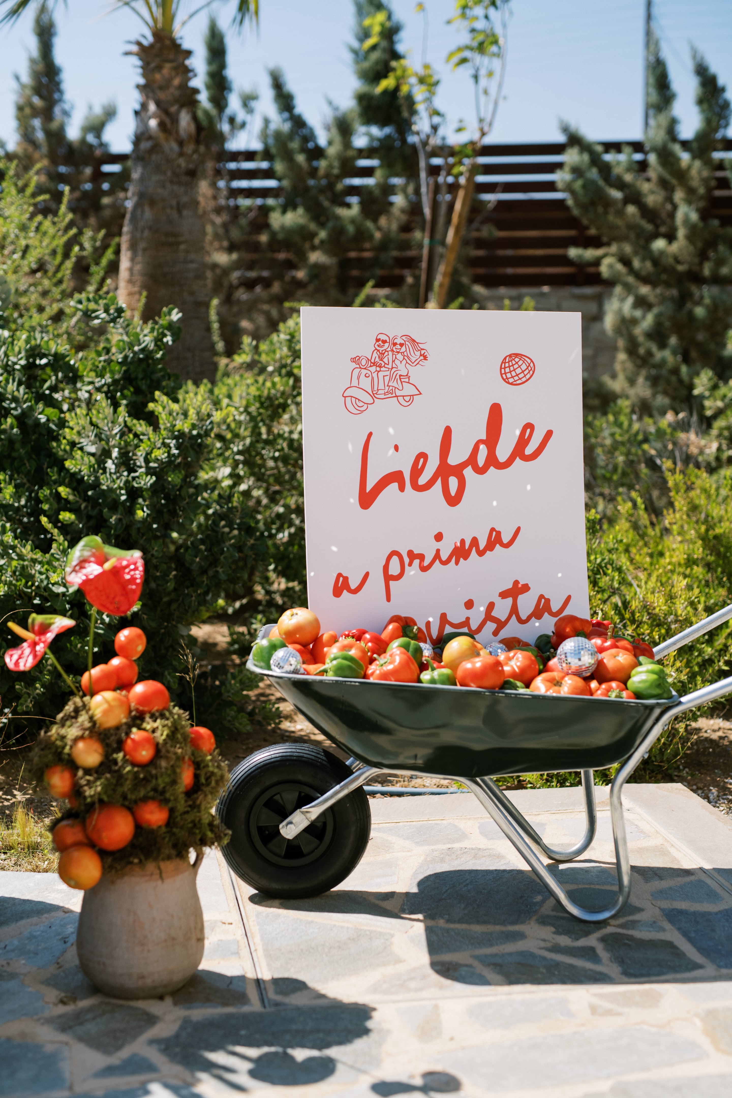A green metal wheelbarrow overflowing with red and green peppers, tomatoes and small silver disco balls stands in a sunny garden, in front of a white sign with red hand lettering reading "Liefde a prima vista" and a red line drawing of a bride and groom on a scooter. Beside it, a moss-covered pot is topped with red anthurium flowers and round orange tomatoes. This is a playful wedding welcome display at Wine Dark Villas in Greece, with bright red and green tones against the soft shrubbery.