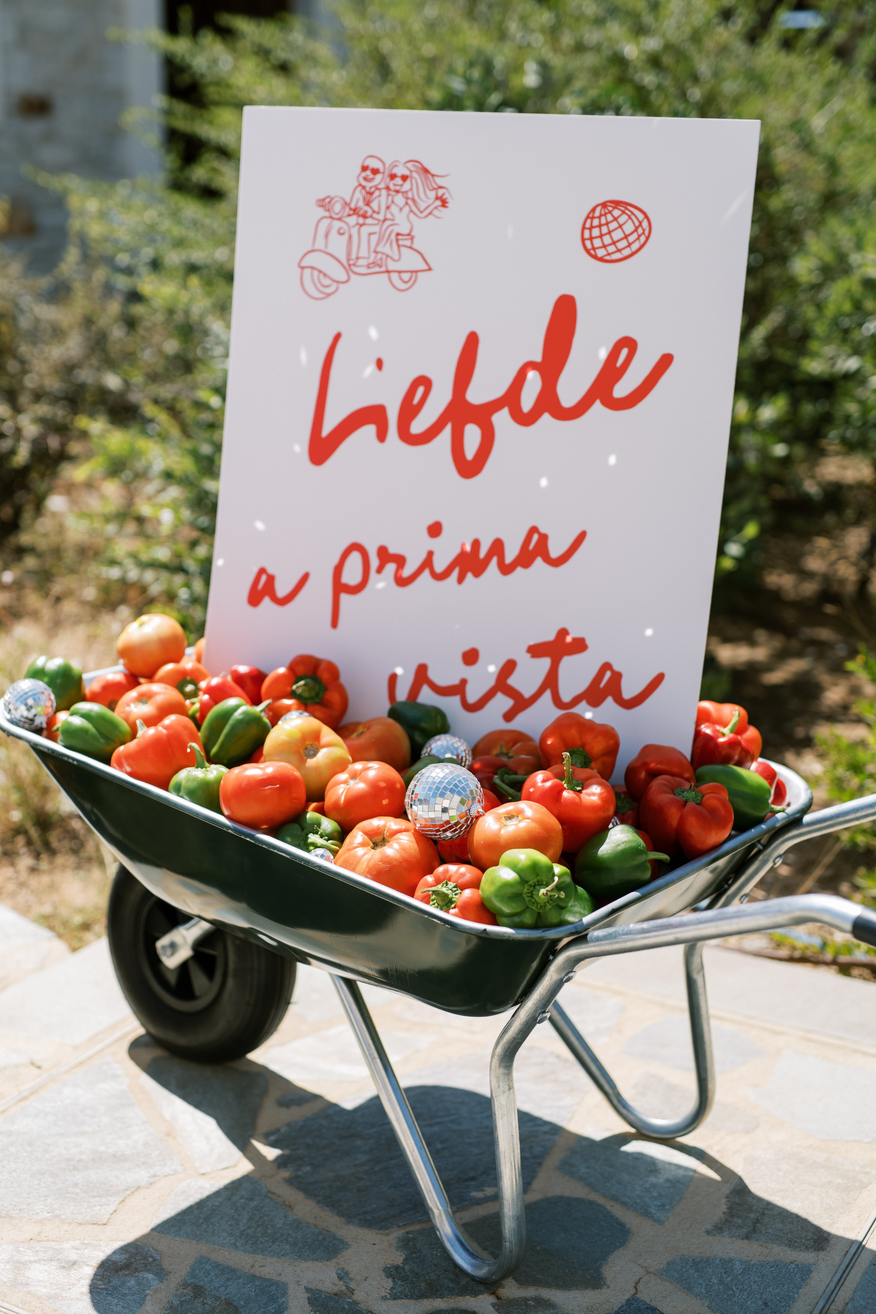A dark green metal wheelbarrow overflowing with red and green bell peppers and ripe tomatoes, with small silver disco mirror balls tucked among them, sits on a stone patio in Greece. Behind it stands a white sign with red handwritten script reading "a prima vista," and sunlight and leafy shade fall across the scene. This playful wedding styling at Wine Dark Villas in Greece turns produce into a cheerful table display.