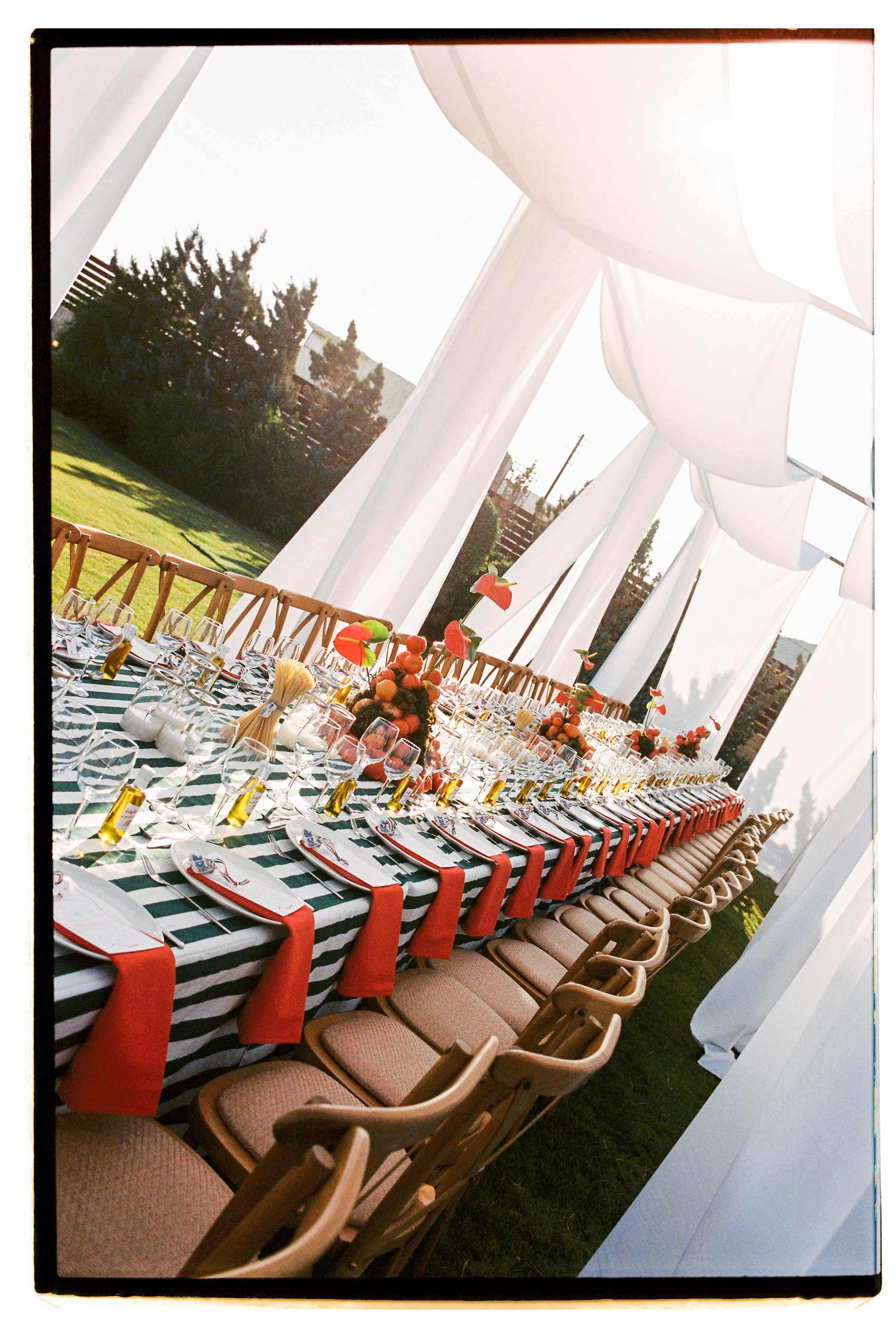 A long banquet table under white draped marquee canopies, dressed with a green and white striped tablecloth and red napkins laid across each place setting. Clusters of orange fruit and red anthurium blooms rise along the centre, with clear wine glasses and small bottles of olive oil lined up beside the plates. Wood and padded chairs line the side in soft late-afternoon light, at a wedding table flower setting in Greece at Wine Dark Villas.