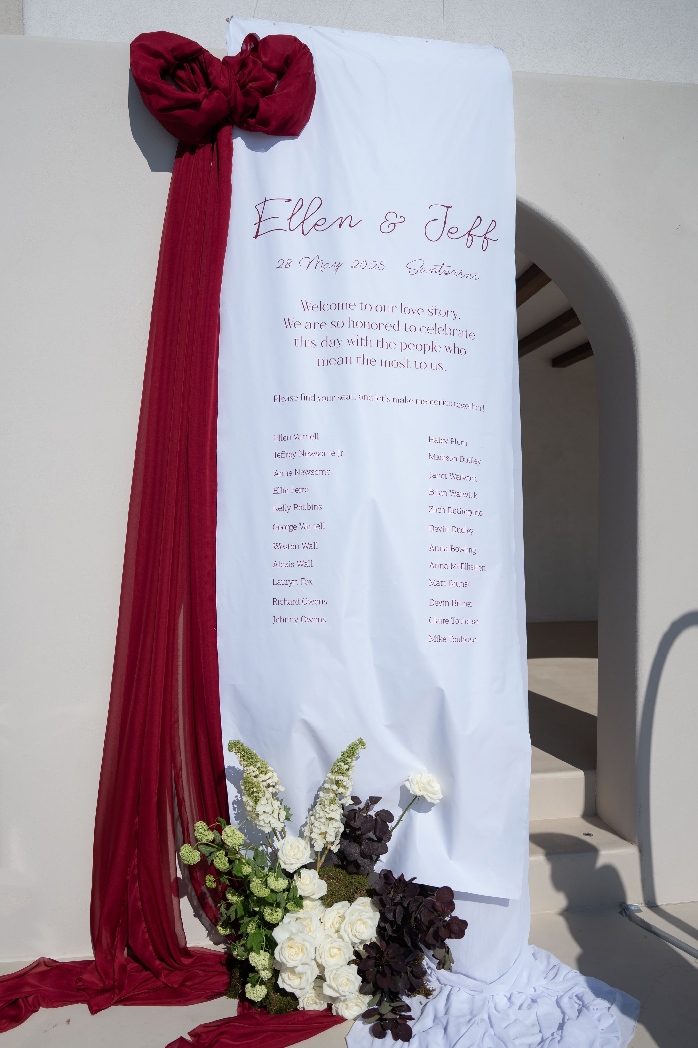 Wedding flowers at Islet in Santorini, Greece: a cascade of white garden roses, pale green viburnum and tall white delphinium spikes sits at the foot of a long white fabric panel printed with guest names. Deep burgundy chiffon falls down one side, and dark plum foliage, possibly oxalis or aeonium, adds drama among the blooms and soft moss. The bright, sunlit whitewashed wall and steps behind create a calm, elegant mood.