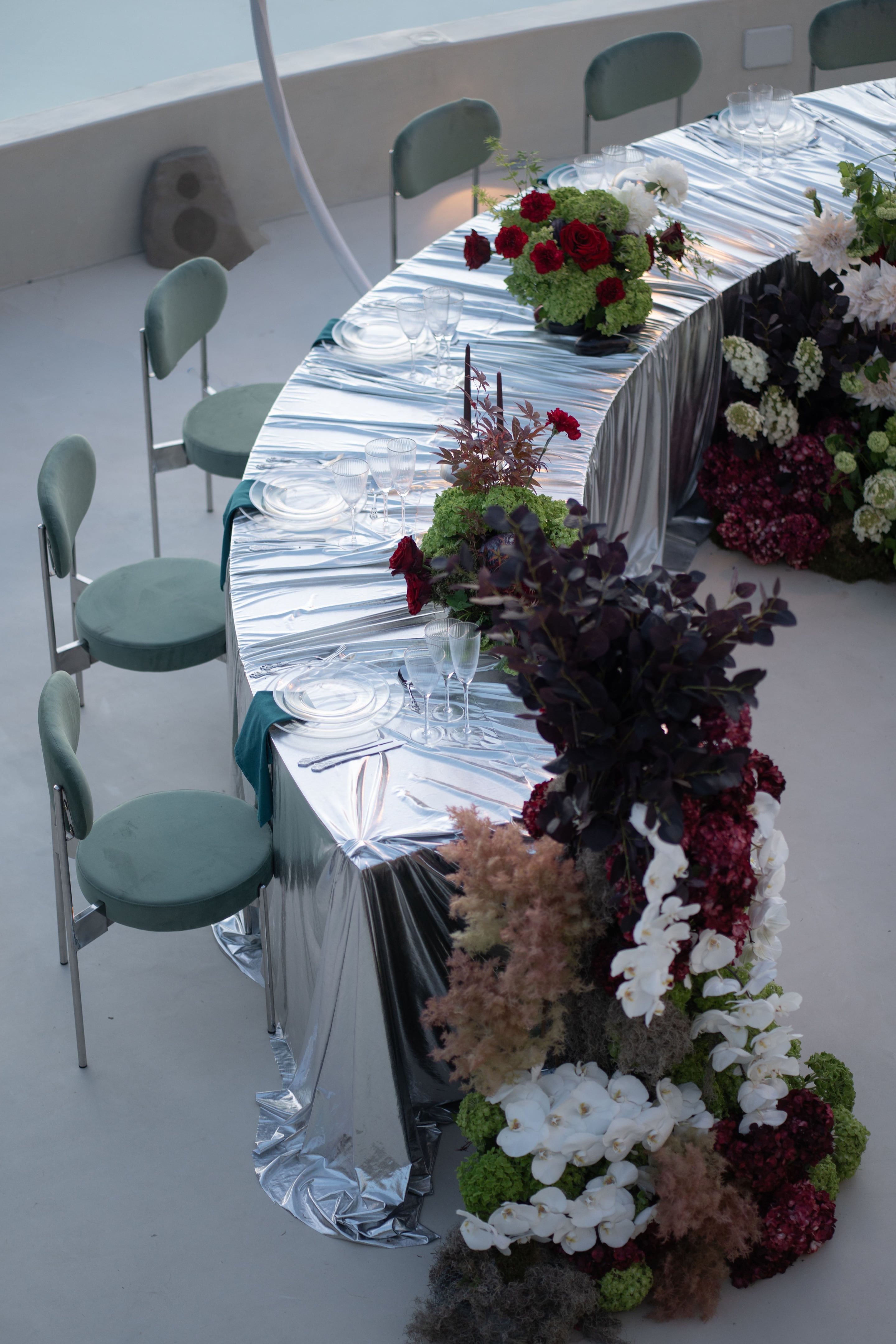 A curved head table draped in rumpled silver lamé, set with clear glass plates, stemmed glassware and teal napkins, with sage-green velvet chairs down one side. Along the edge runs a lush arrangement of deep burgundy roses, dark plum foliage, cream hydrangeas and white orchids, with a low green-and-red centrepiece of roses and dark leaves in the middle. This wedding table flower design is at Islet in Santorini, Greece, and the soft overhead light gives it a moody, elegant feel.