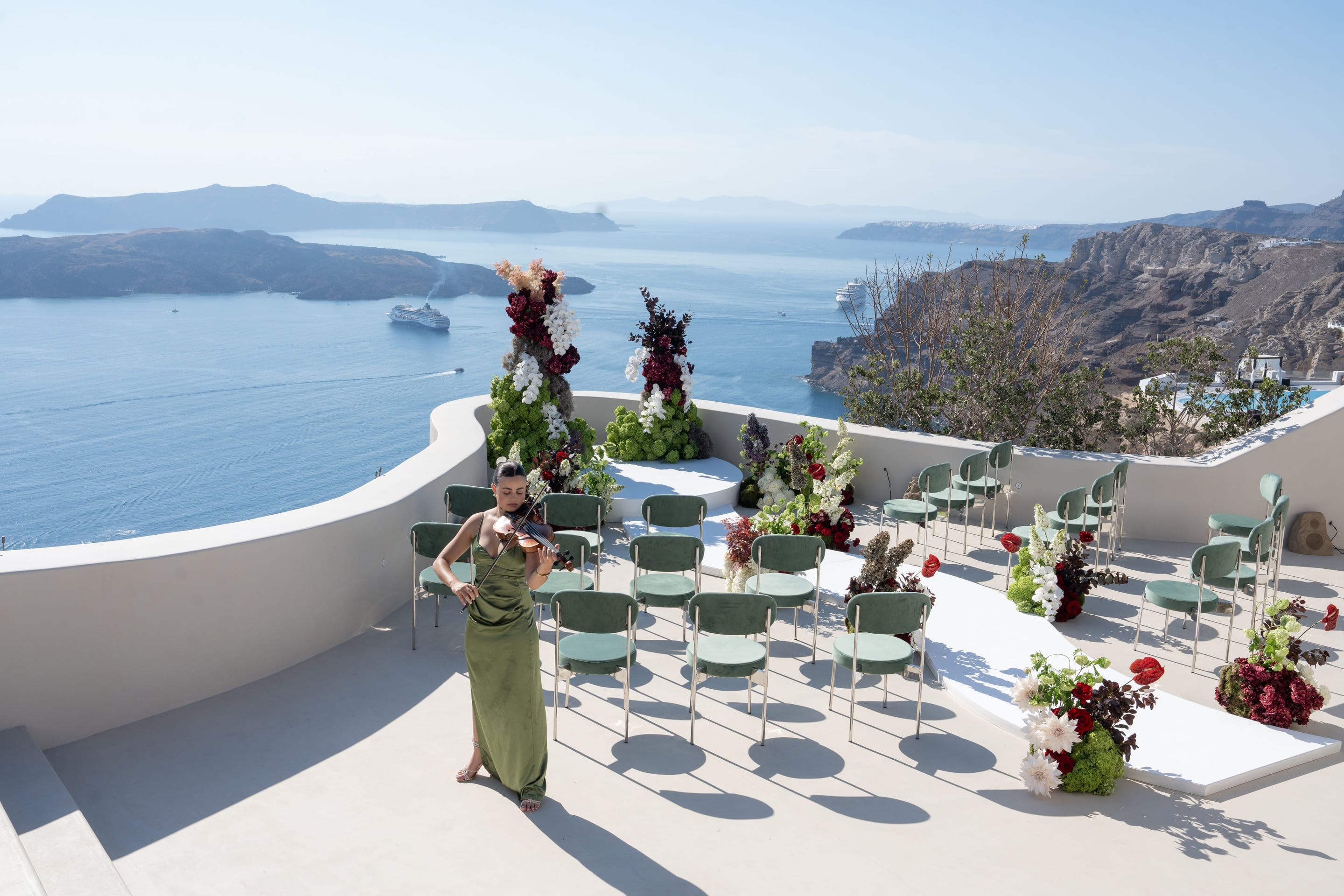 A violinist in a long olive-green satin gown plays on a whitewashed clifftop terrace overlooking the caldera at Islet Santorini, Greece, where a wedding ceremony is set up with rows of sage-green velvet chairs. Tall floral arrangements of white blooms, deep burgundy roses, and anthuriums with dark foliage frame a raised white altar and an aisle, while cruise ships and distant islands drift on the blue sea under a pale, hazy sky.