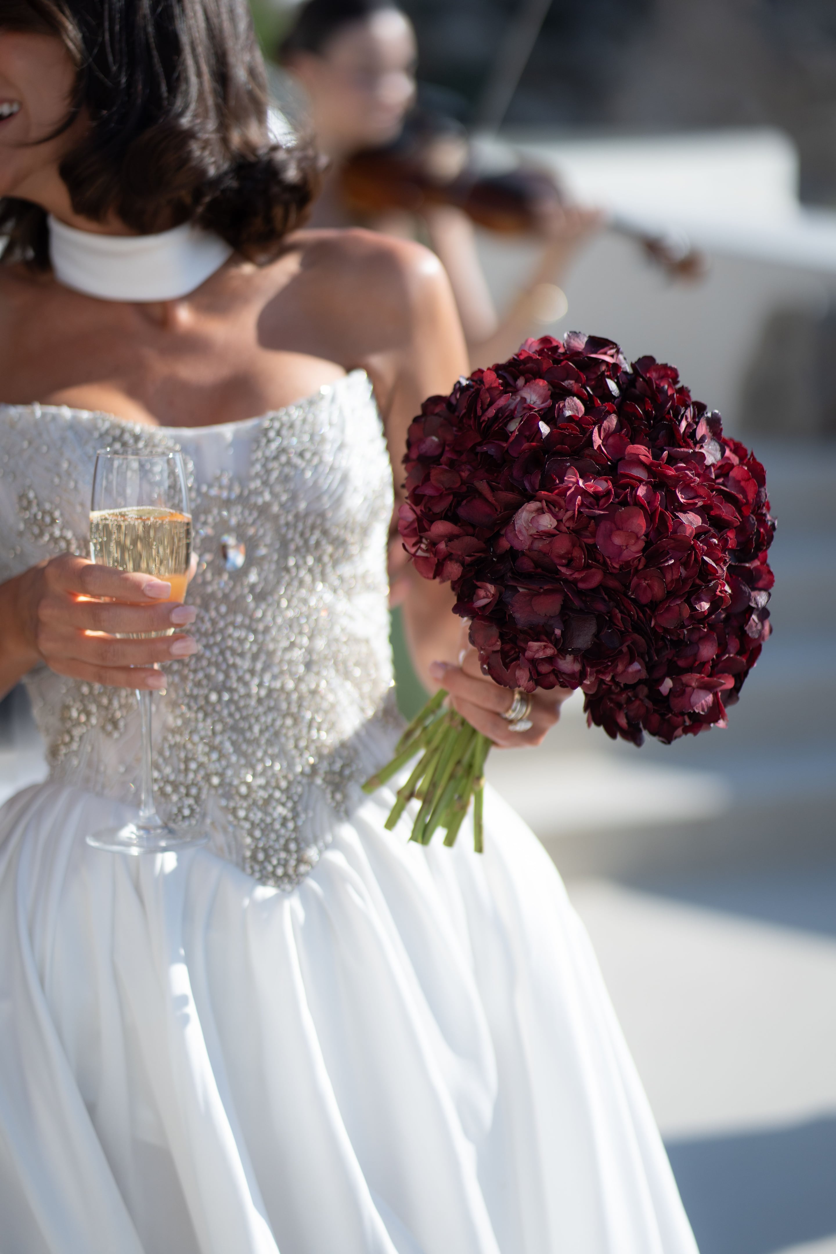 A bride with dark, wavy shoulder-length hair and a white high neck choker holds a flute of champagne in one hand and a dense, rounded bouquet of deep burgundy hydrangea blooms in the other, on a sunlit wedding day at Islet in Santorini, Greece. Her beaded white gown catches the light, and a violinist stands softly out of focus behind her against pale white walls.