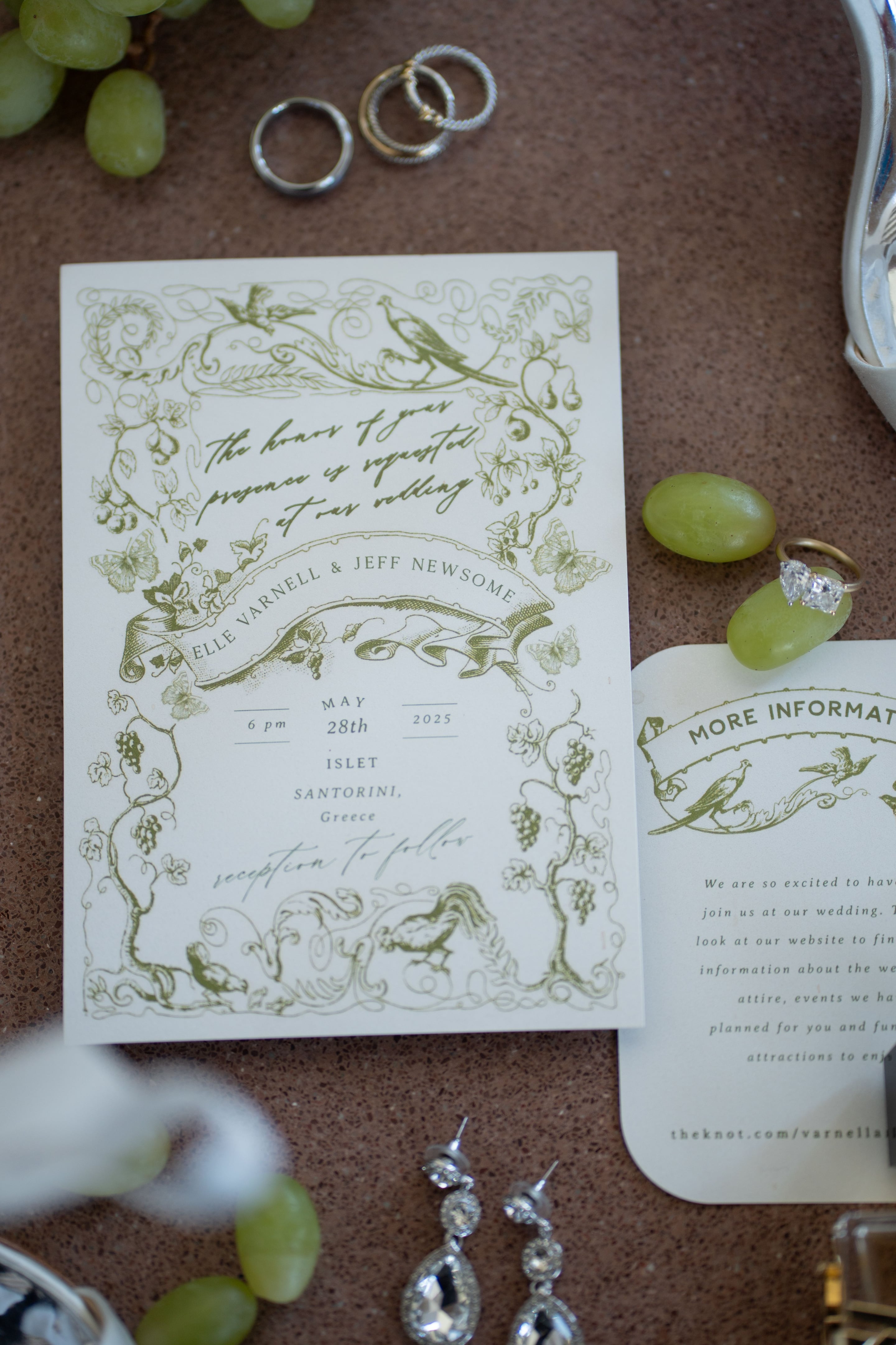 A flat-lay of a cream wedding invitation printed in olive green with an ornate border of grape vines, leaves, butterflies and birds, and a banner reading Elle Varnell and Jeff Newsome. The invitation gives the date of May 28th, 2025, at 6 pm at Islet in Santorini, Greece, with the reception to follow. Beside it sit a green grape, a gold ring with a pair of pale diamonds, and a smaller "More Information" card.
