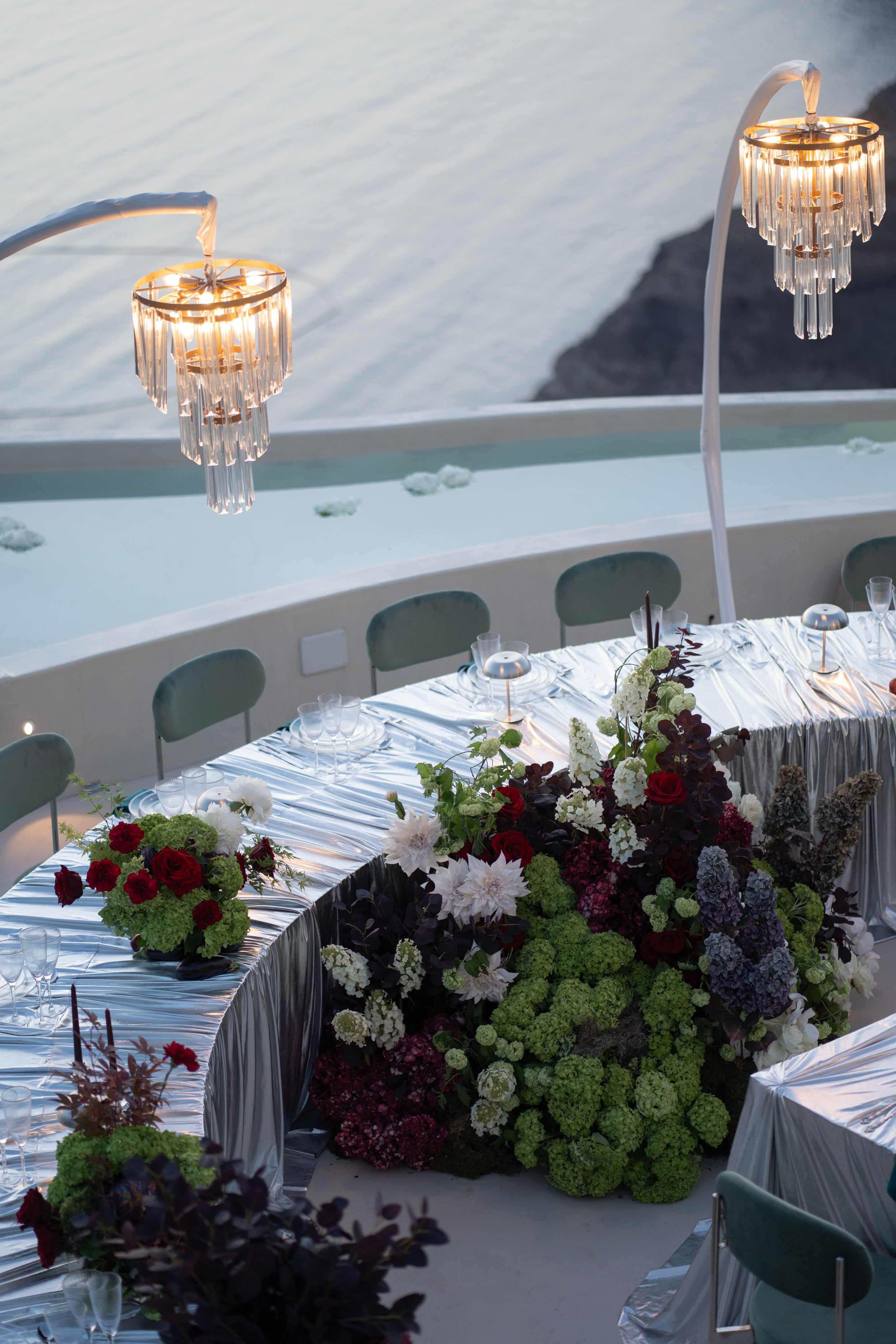 A long curved wedding table at Islet Santorini in Greece, draped in shimmering silver fabric and set with clear glassware and small brass-topped lamps. Along its length runs a lush arrangement of green hydrangeas, deep red garden roses, white dahlias and white snowball blooms, with dark plum foliage and spires of blue-grey flowers threaded through. A smaller red-and-green rose and hydrangea centrepiece sits at the far end, with a pool and white chair-backed seating behind it.
