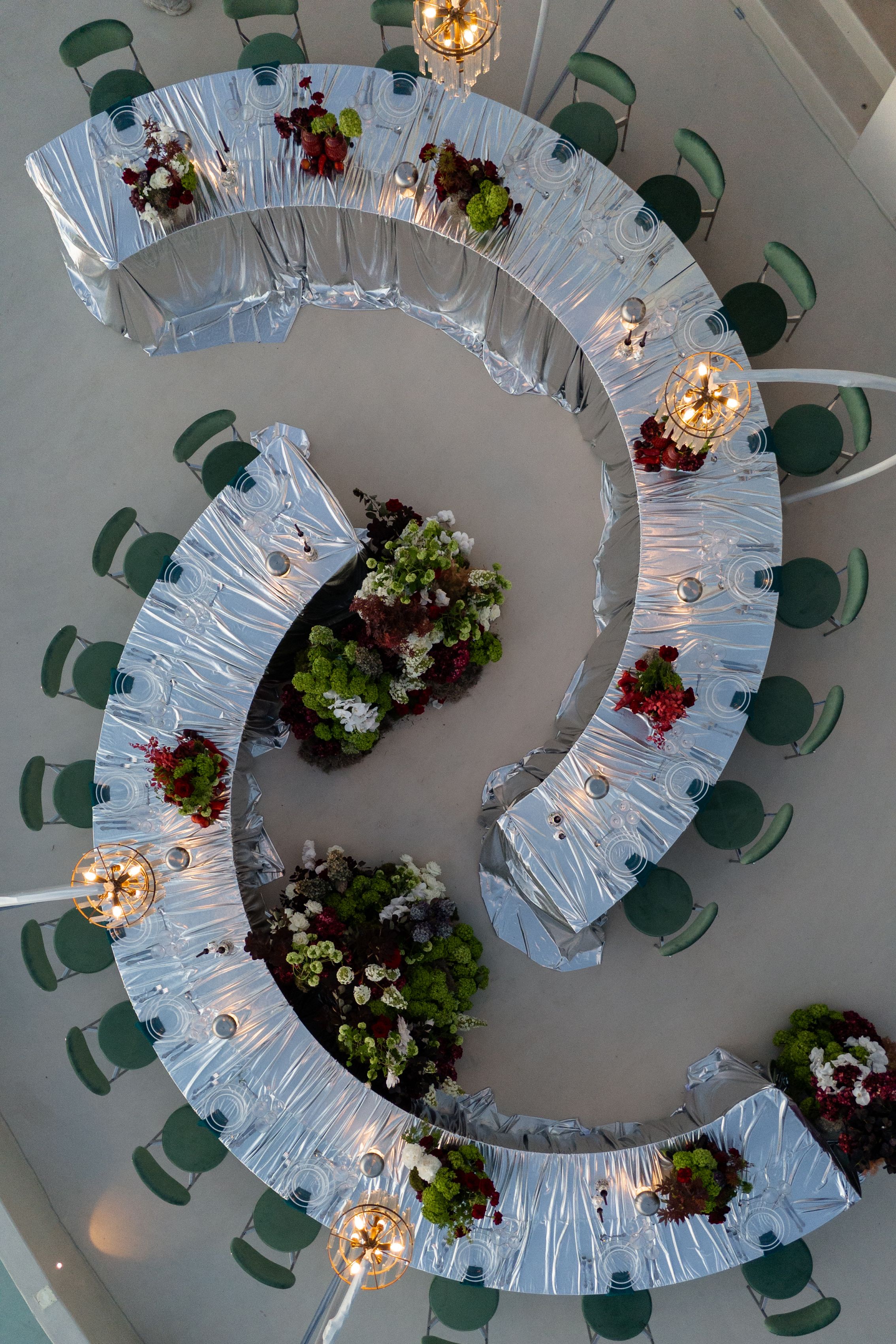 Aerial view of curved silver-draped banquet tables at Islet Santorini in Greece, set for a wedding reception. Along the tables sit clusters of green hydrangea-like blooms, white flowers and deep burgundy foliage, with small red roses and dark berry-toned sprigs at intervals, and little glowing candles and crystal glasses catching the light. Green-cushioned chairs line the outer edges, and warm pendant lights hang above.