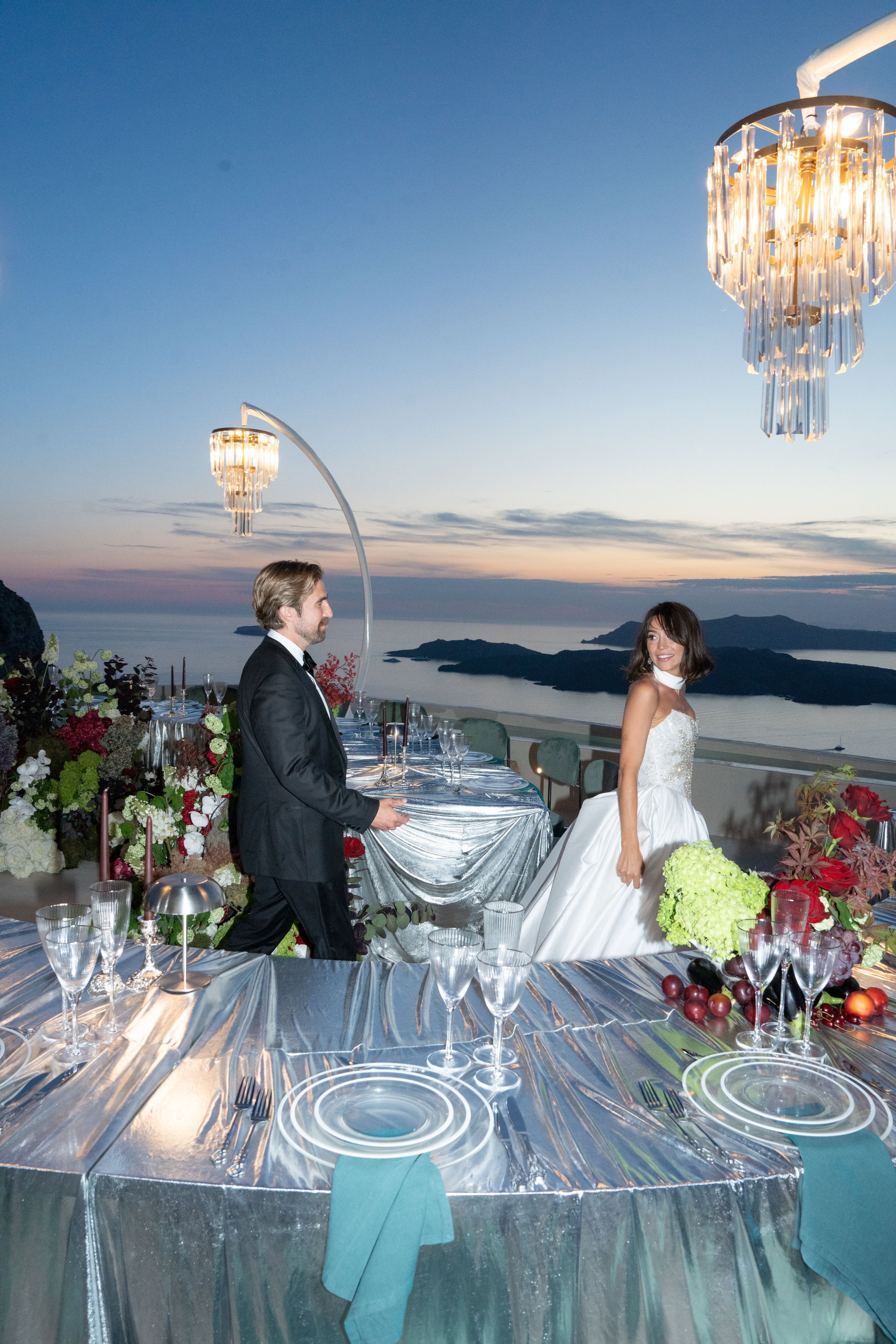 A bride in a strapless white ballgown with a sparkling bodice and a soft white neck tie glances over her shoulder with a smile, while a groom in a black tuxedo and bow tie stands beside a long reflective silver table on a clifftop terrace at Islet in Santorini, Greece. Lush centrepieces of white hydrangeas, red roses, dark foliage and tall burgundy candles line the table, with crystal chandeliers overhead and the sea and dark islands glowing under a pastel sunset behind them. This is a wedding table styled with flowers in Greece.