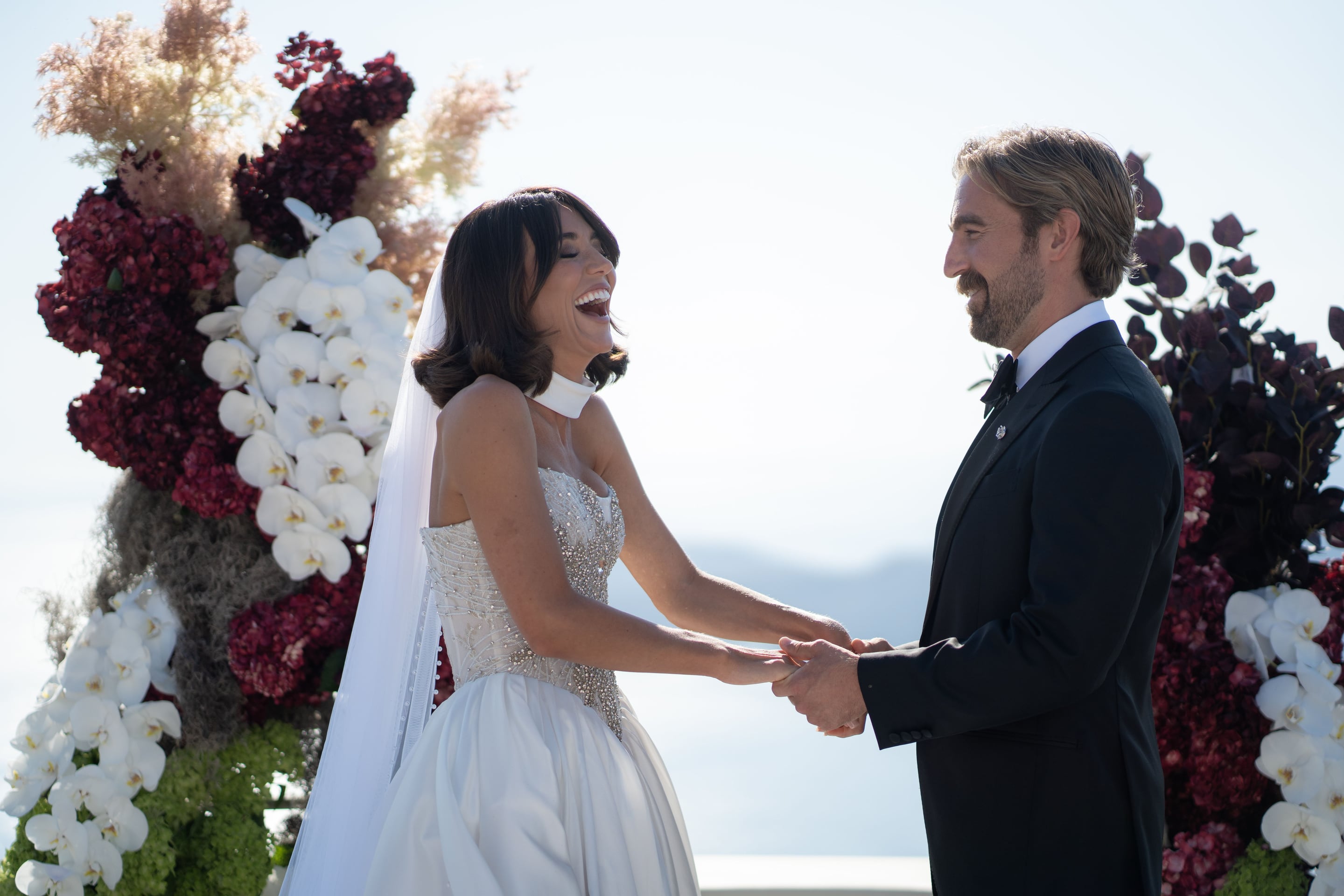 A bride with dark bobbed hair and a sheer veil laughs joyfully as she holds the hands of her smiling groom, who wears a black tuxedo and bow tie, during a wedding ceremony at Islet in Santorini, Greece. Behind them stands a tall arch of deep burgundy hydrangeas, pale plumes of dried grass, and cascading white phalaenopsis orchids, set against a bright, hazy sky over the sea.